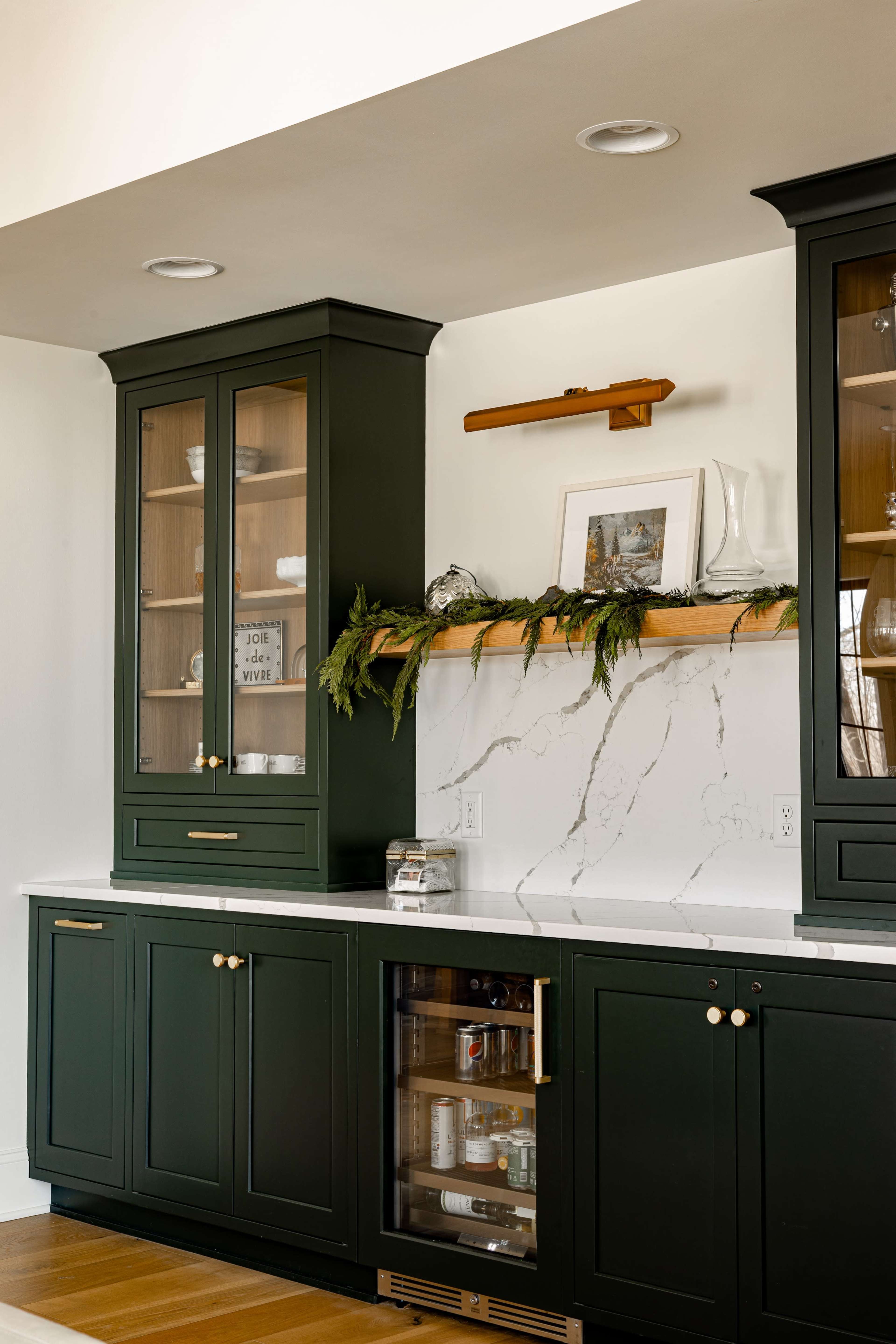 The image shows a modern kitchen corner featuring dark green cabinets, a marble backsplash, and a wooden shelf adorned with greenery and decorative items.