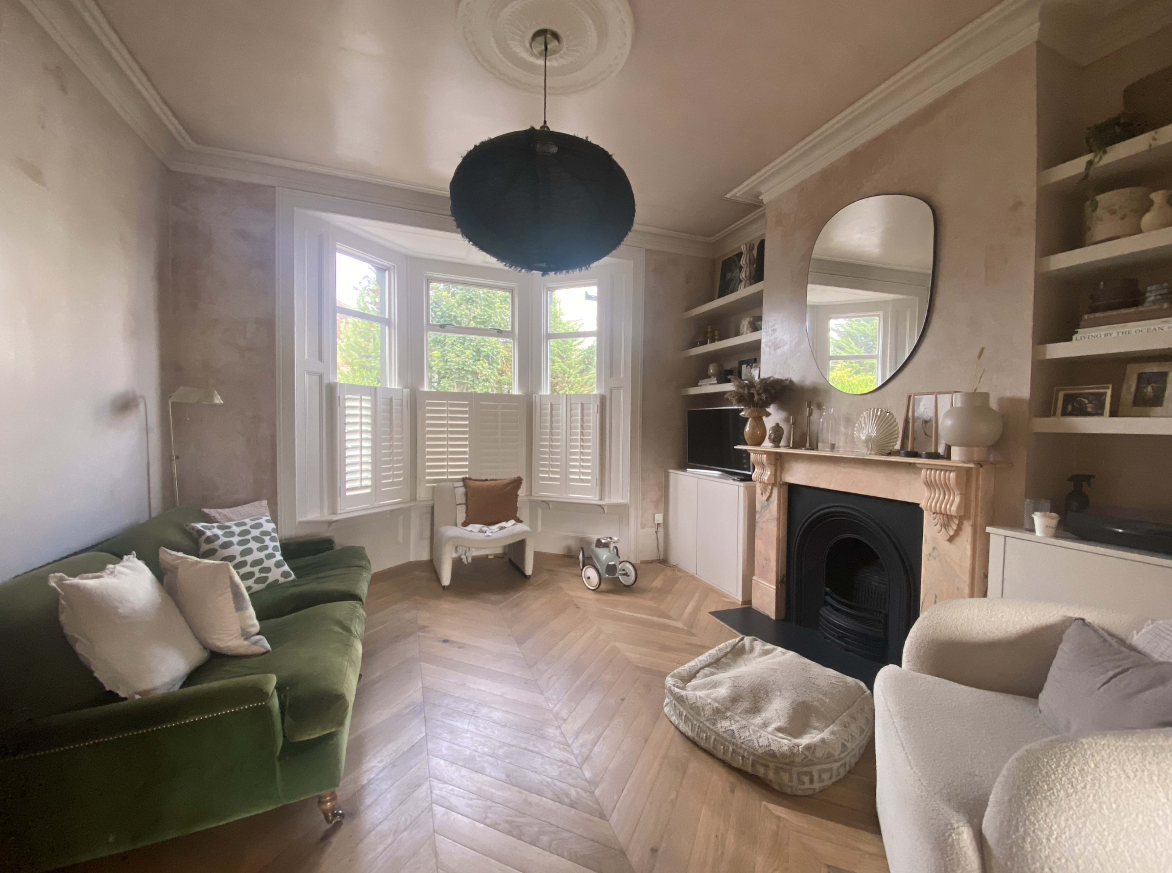 The living room features a green velvet sofa, a white armchair, a fireplace with a mirror above it, and large windows with shuttered blinds.