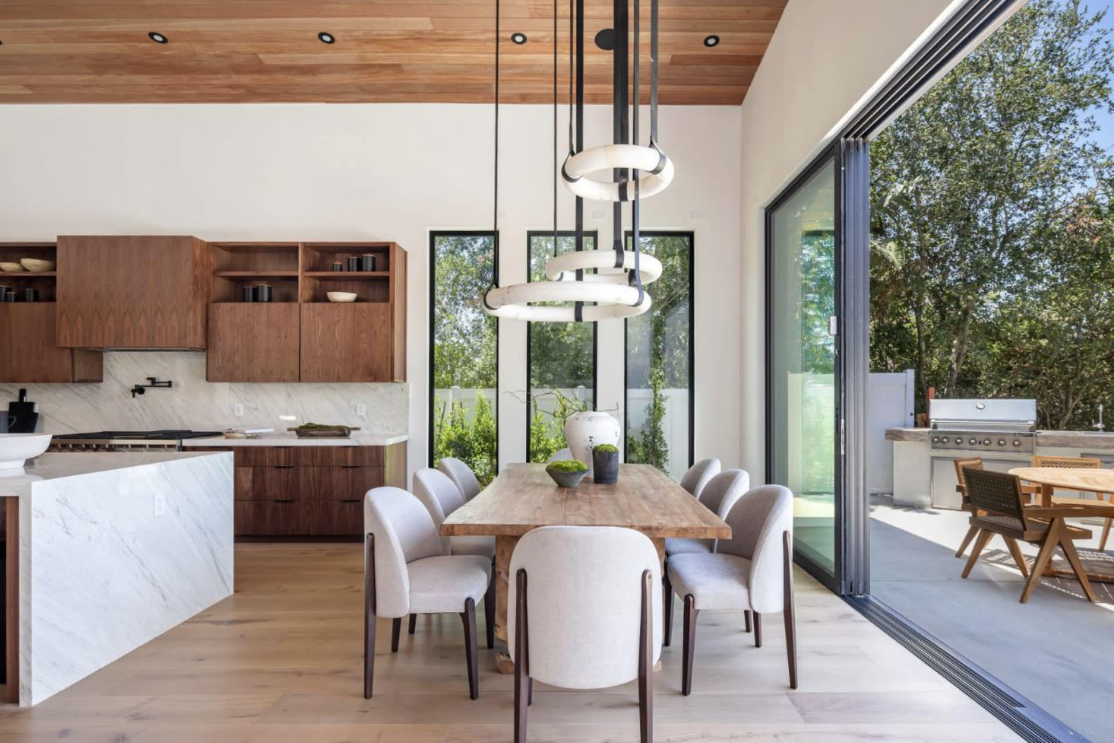 The image shows a modern dining area with a wooden table surrounded by beige chairs, adjacent to a sleek kitchen and large windows overlooking an outdoor space.