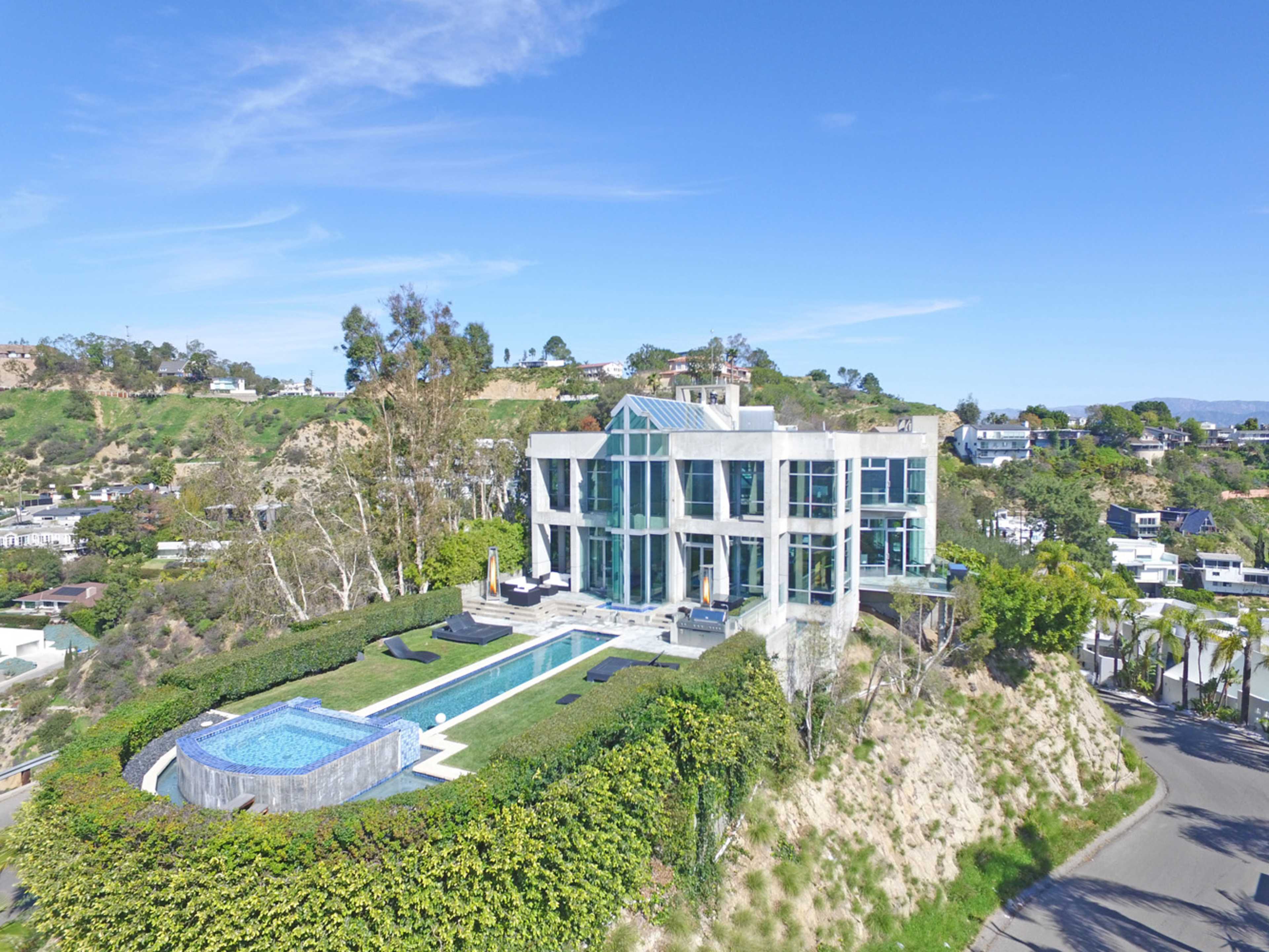 The image shows a modern multi-story house with large glass windows, a rectangular swimming pool, and landscaped greenery on a hillside.