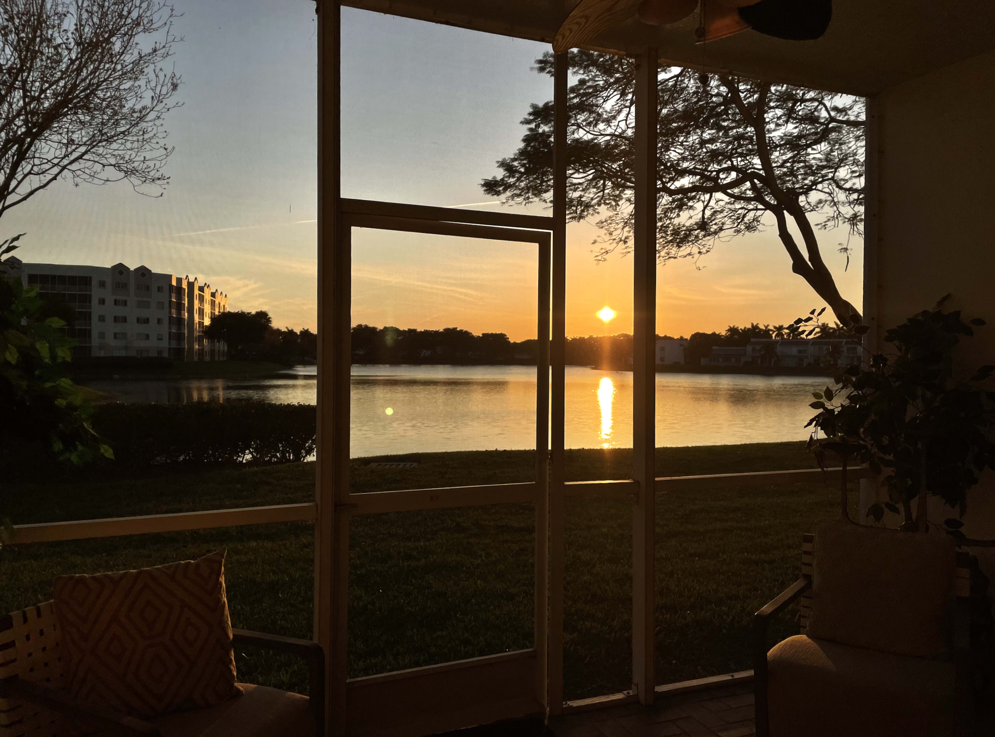 The image shows a sunset view over a lake, framed by a screened porch with a chair and greenery visible.