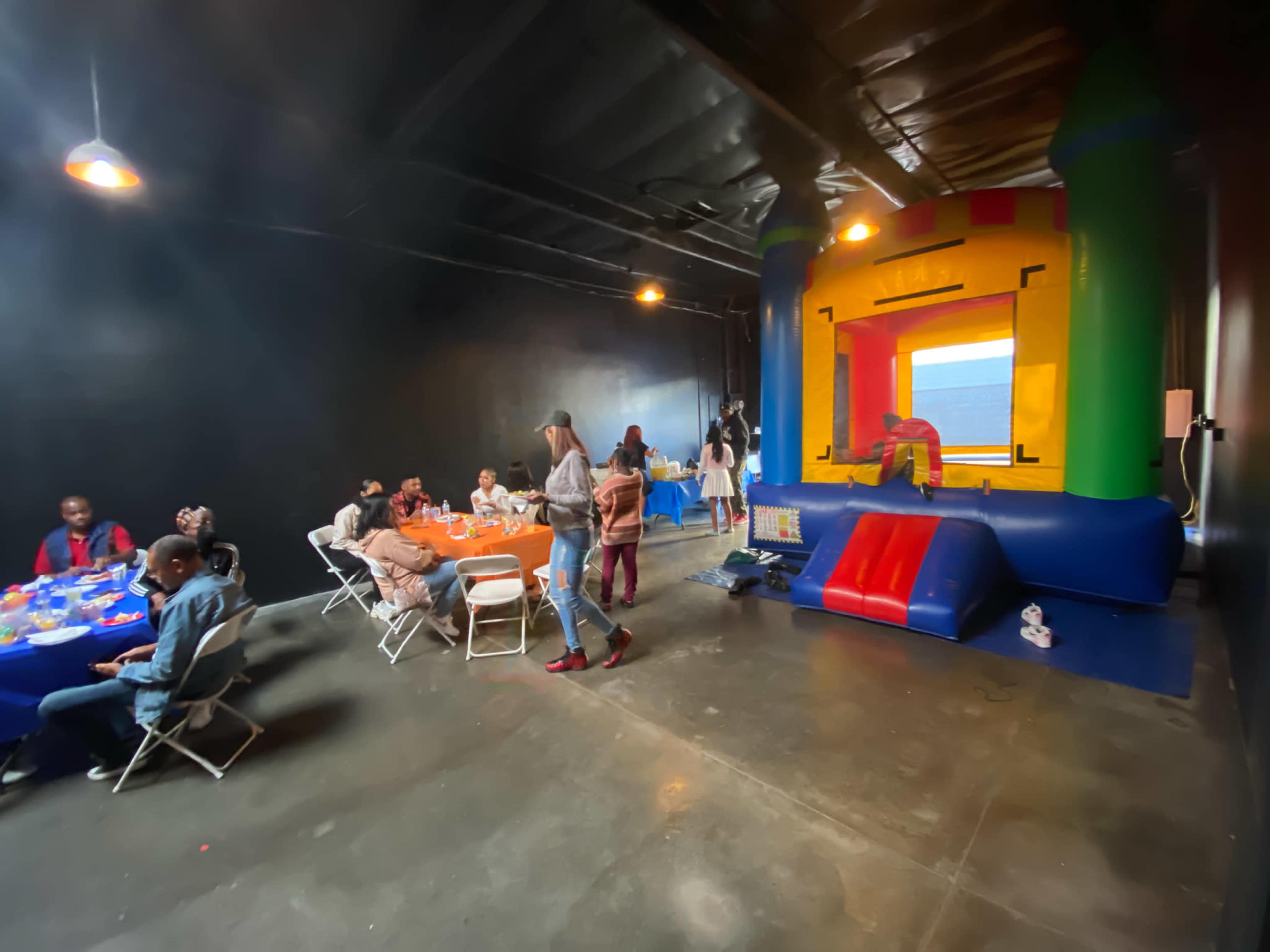 A group of people sits at tables while a colorful inflatable bounce house stands in the corner of a dimly lit room.
