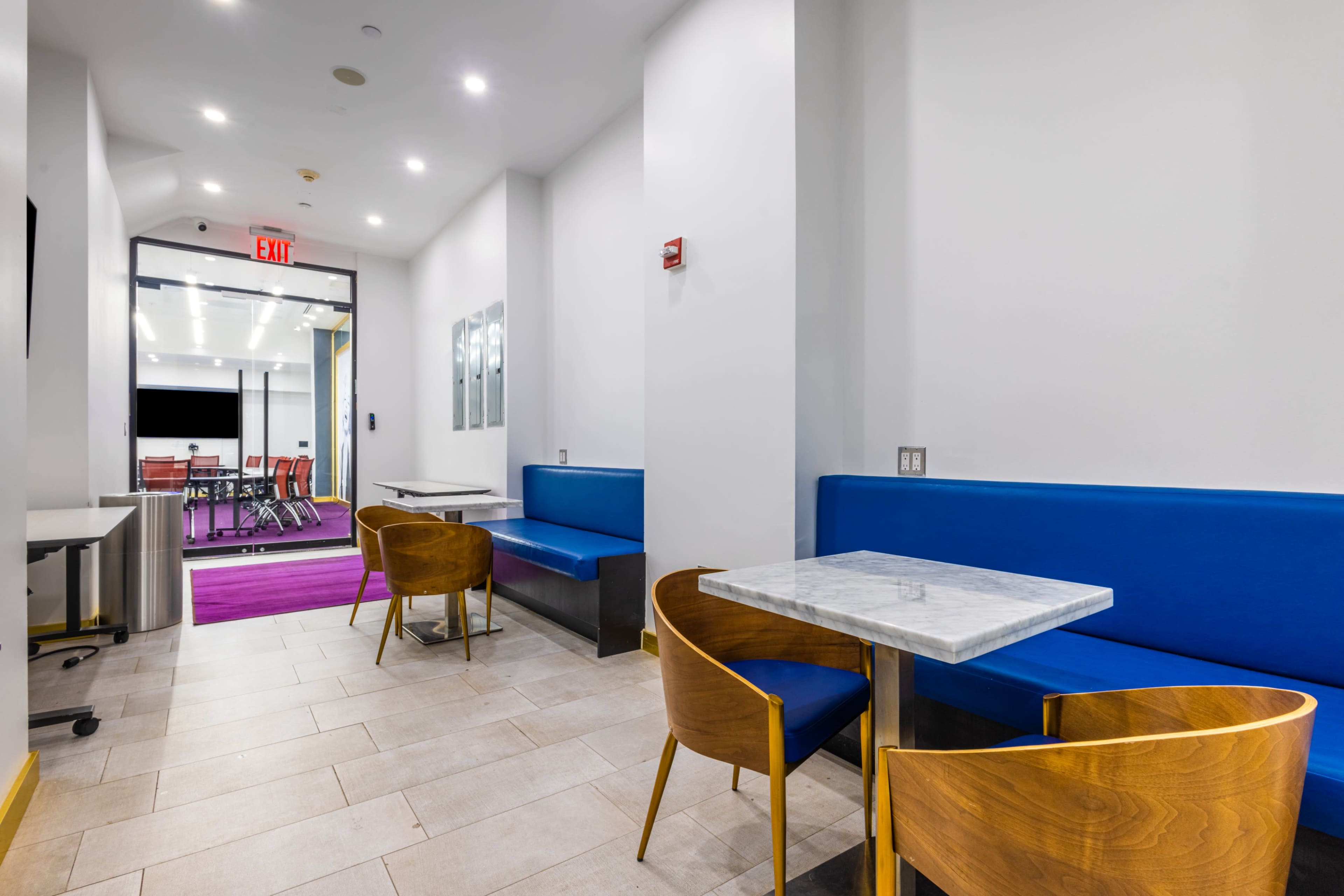 The image shows a modern hallway with blue seating and wooden chairs, leading to a room with a purple carpet and conference setup.