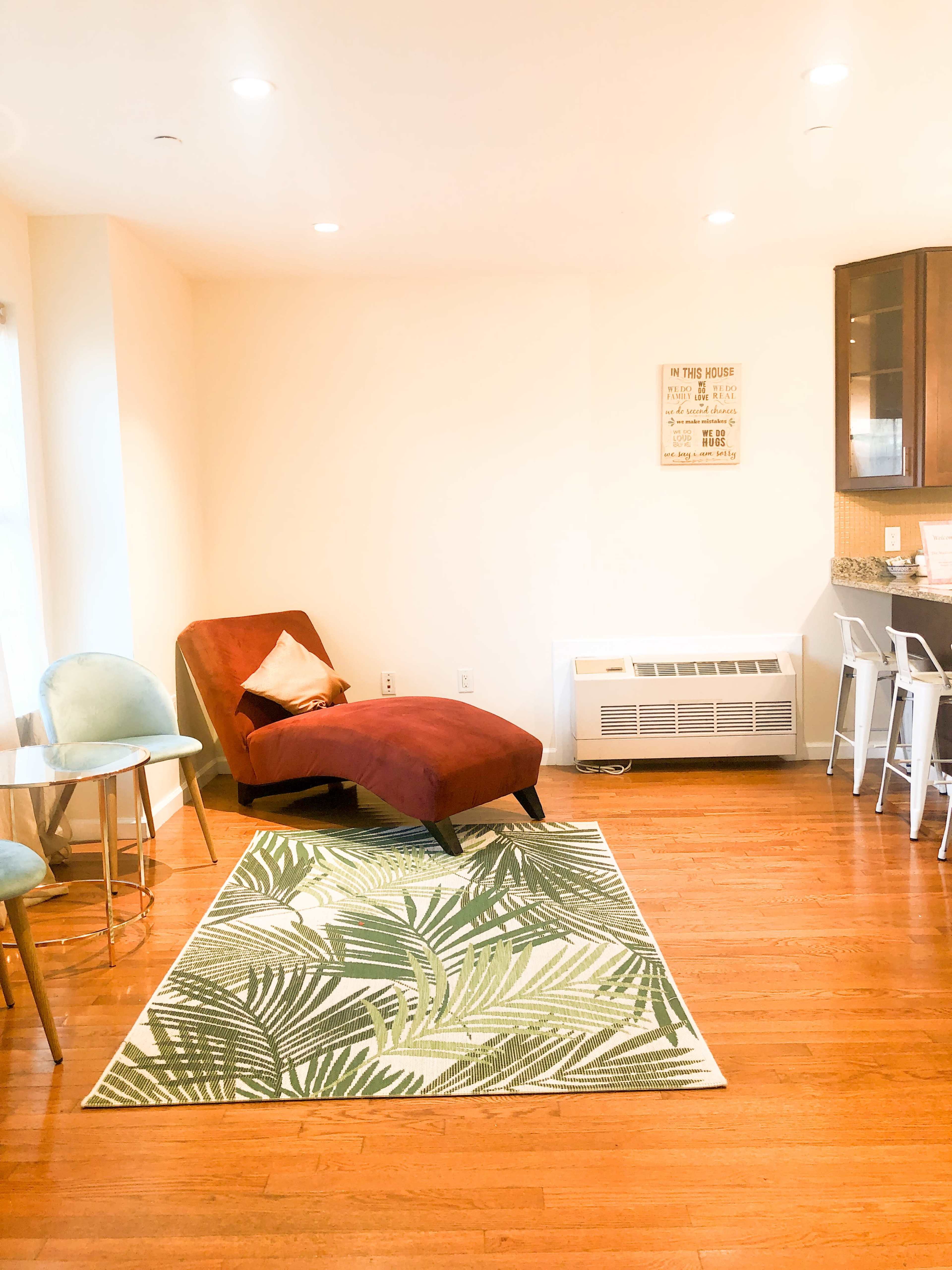 The image shows a room with a red armchair, a green leaf-patterned rug, a small dining table with blue chairs, and a kitchen area in the background.