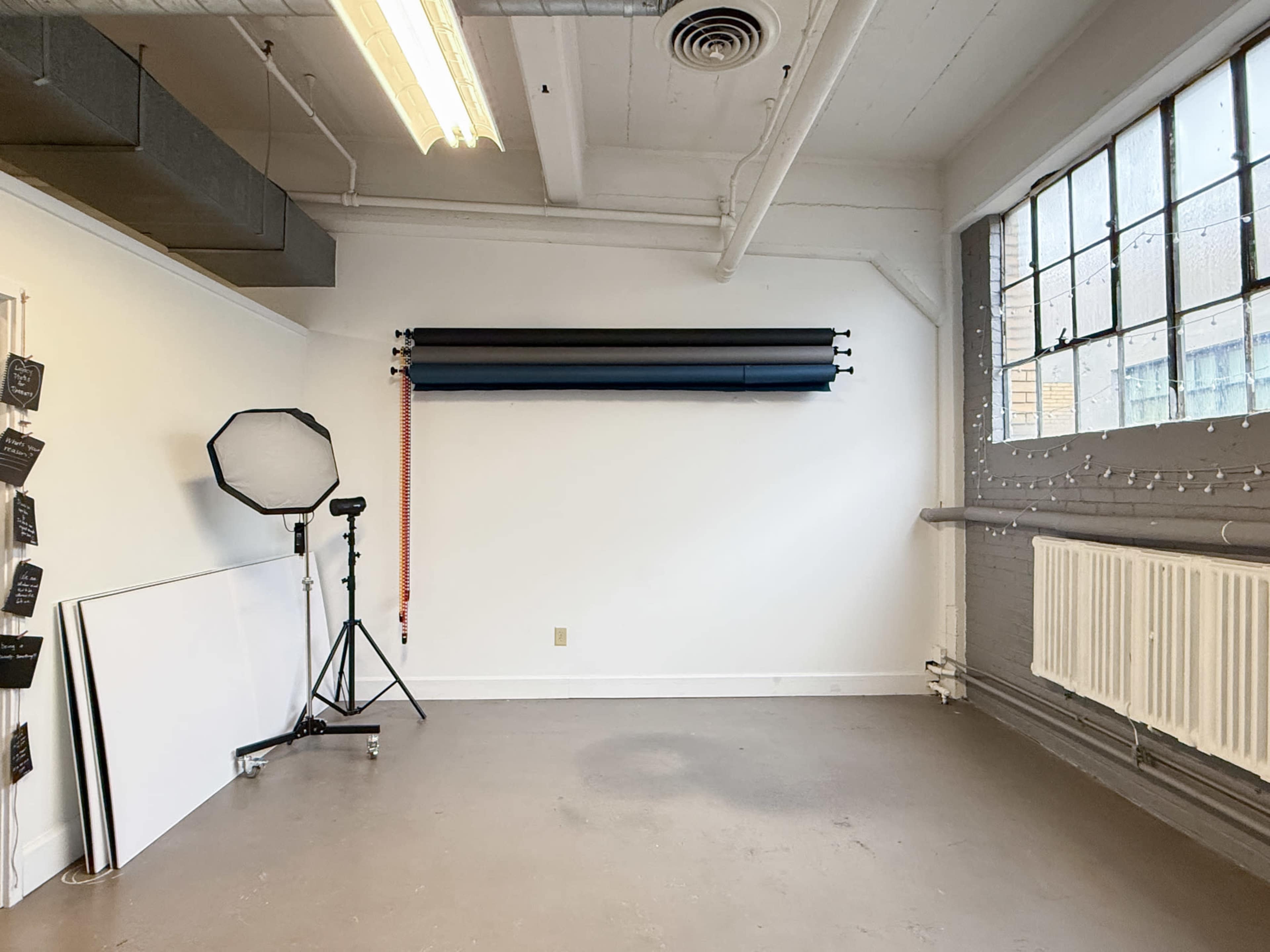 The image shows a spacious, well-lit studio with a wall-mounted backdrop system and a photography setup, including a tripod and a light source.
