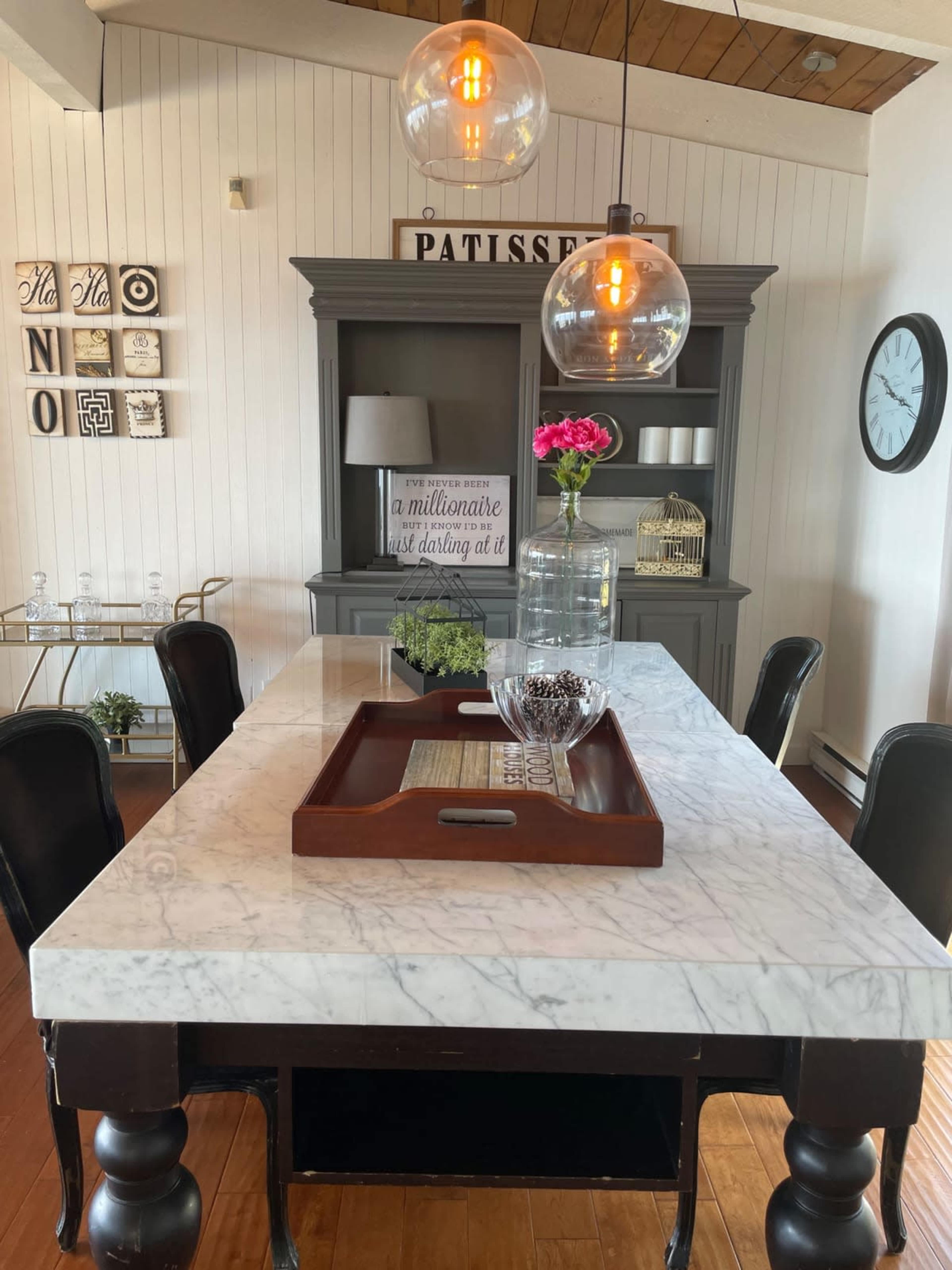 A dining table with a marble top is set with a wooden tray, a glass vase with flowers, and decorative items, surrounded by black chairs in a well-lit room featuring a grey cabinet and wall decor.