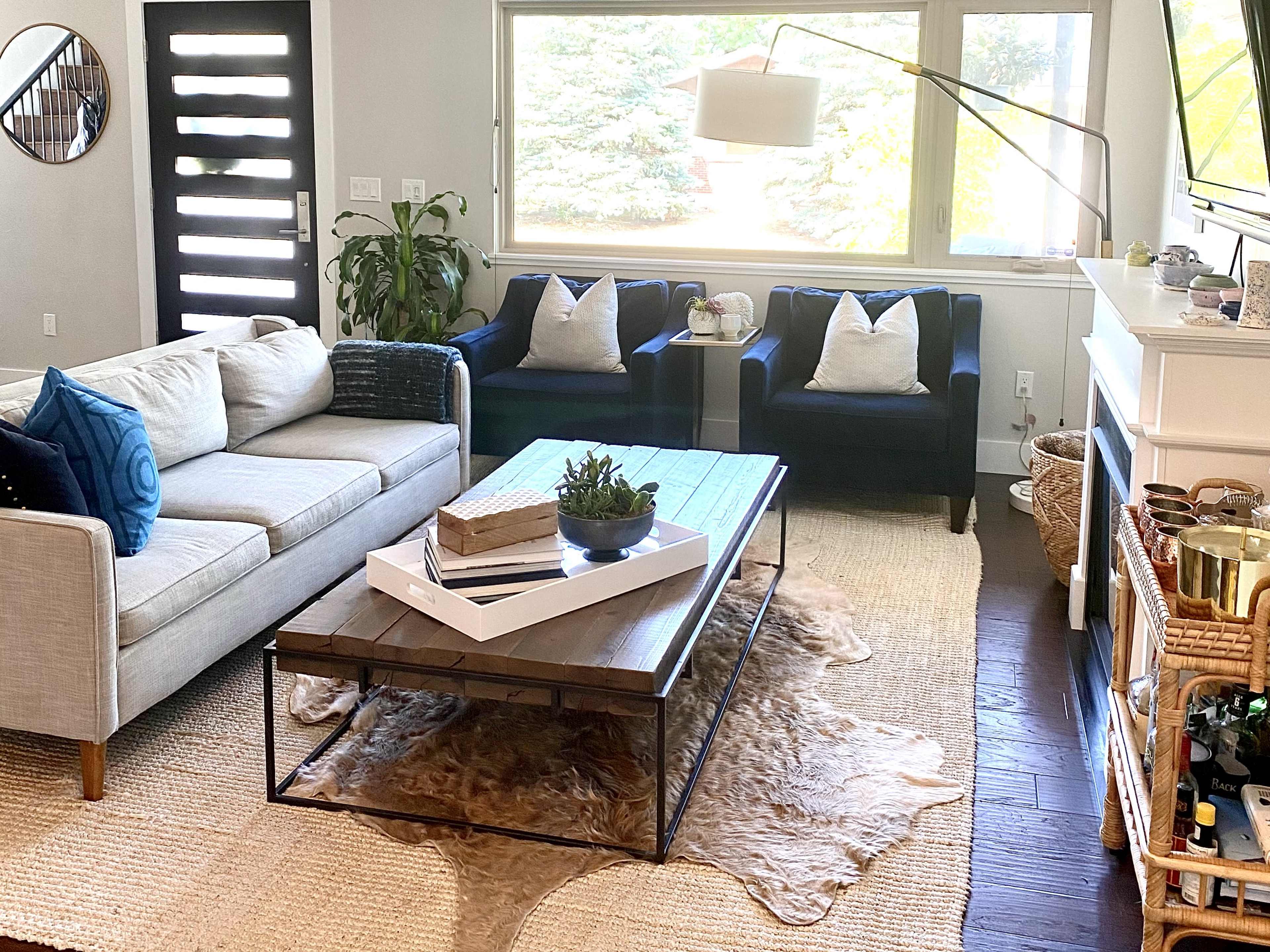 A modern living room features a gray sofa, two dark armchairs, and a wooden coffee table on a light area rug with a decorative plant and books atop it.