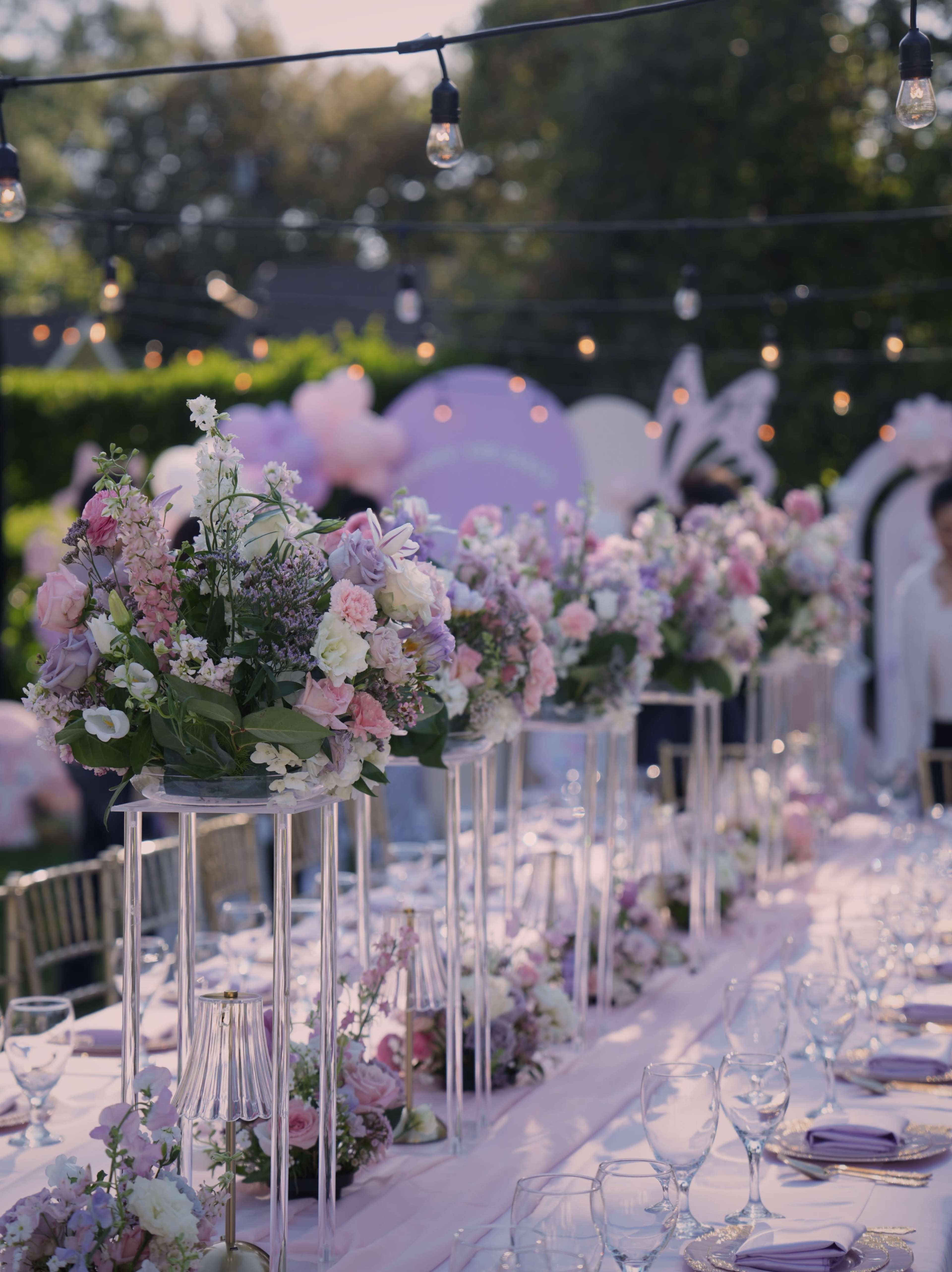 The image shows a long banquet table adorned with floral centerpieces, set outdoors under string lights.