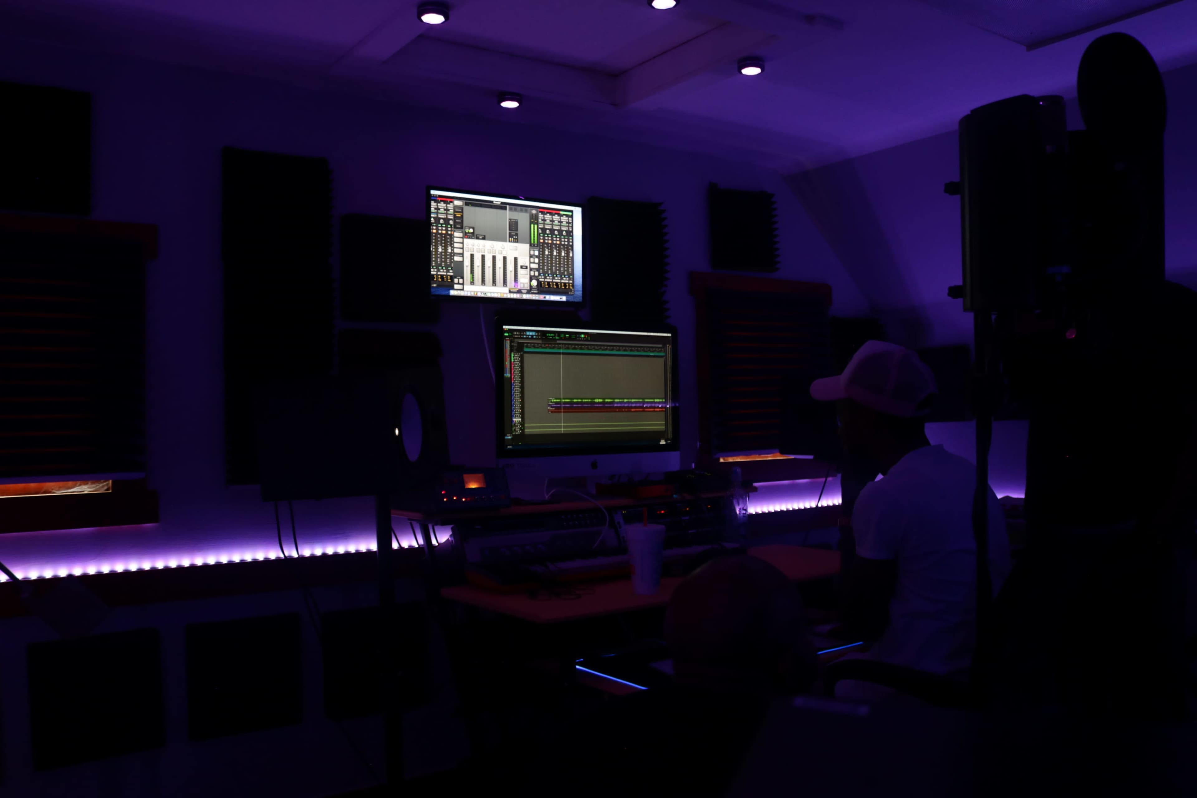 A person sits in a music studio, working on a computer with sound-editing software illuminated by dim purple lights.