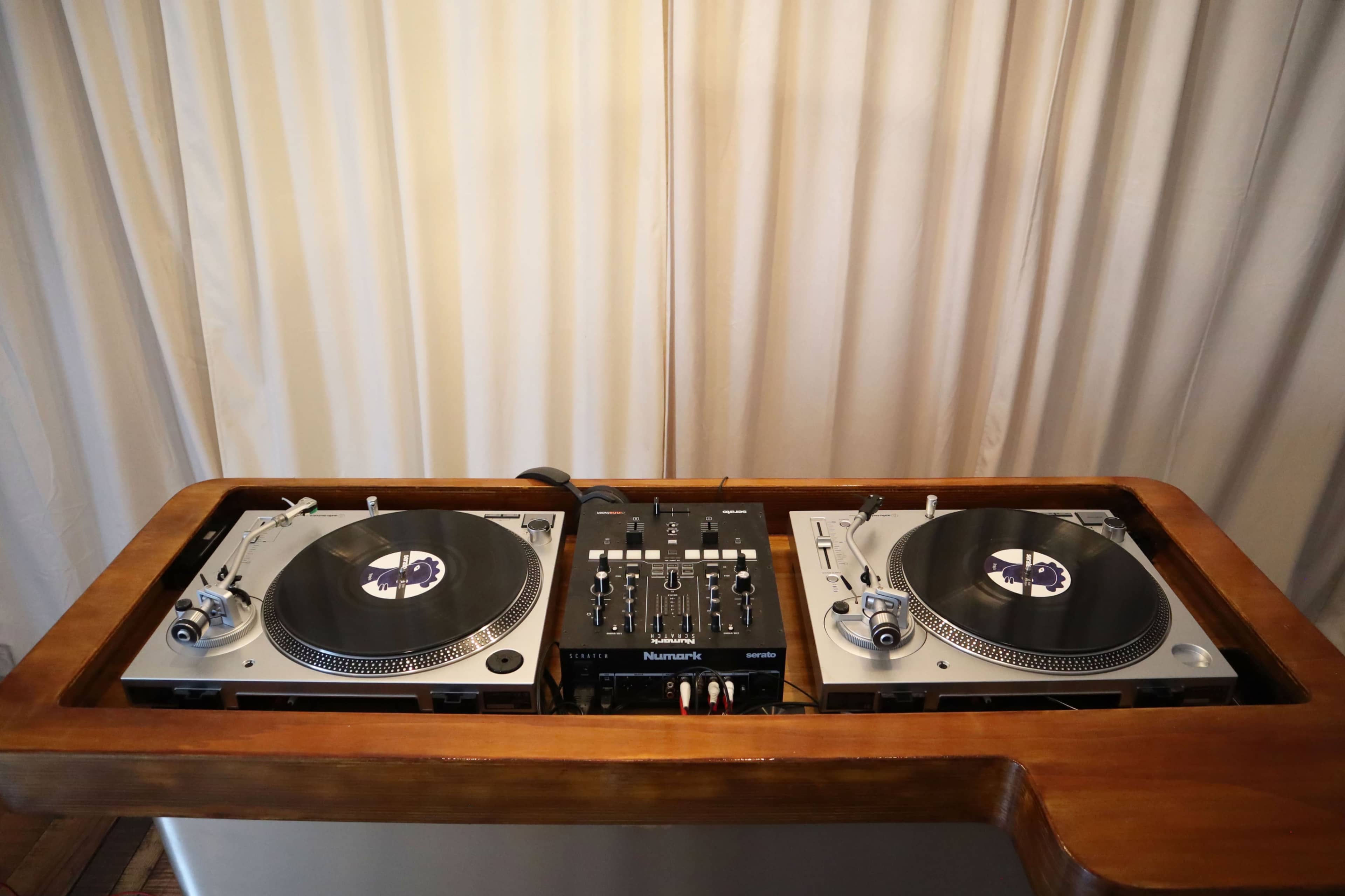 A DJ setup features two turntables and a mixer on a wooden surface with a white curtain in the background.