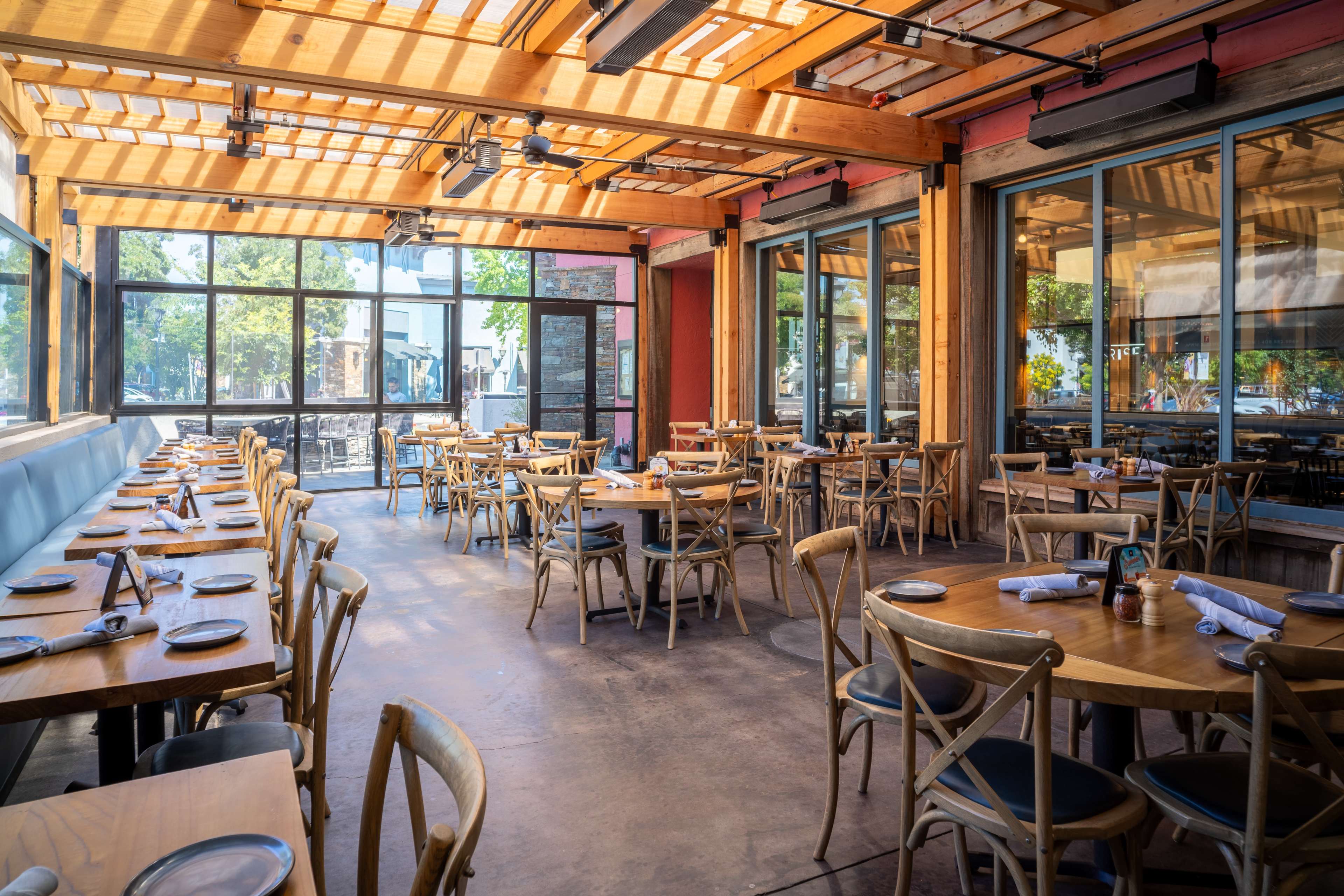 The image shows a bright, modern restaurant with wooden tables and chairs arranged in a spacious, open area featuring large windows and a wooden ceiling.