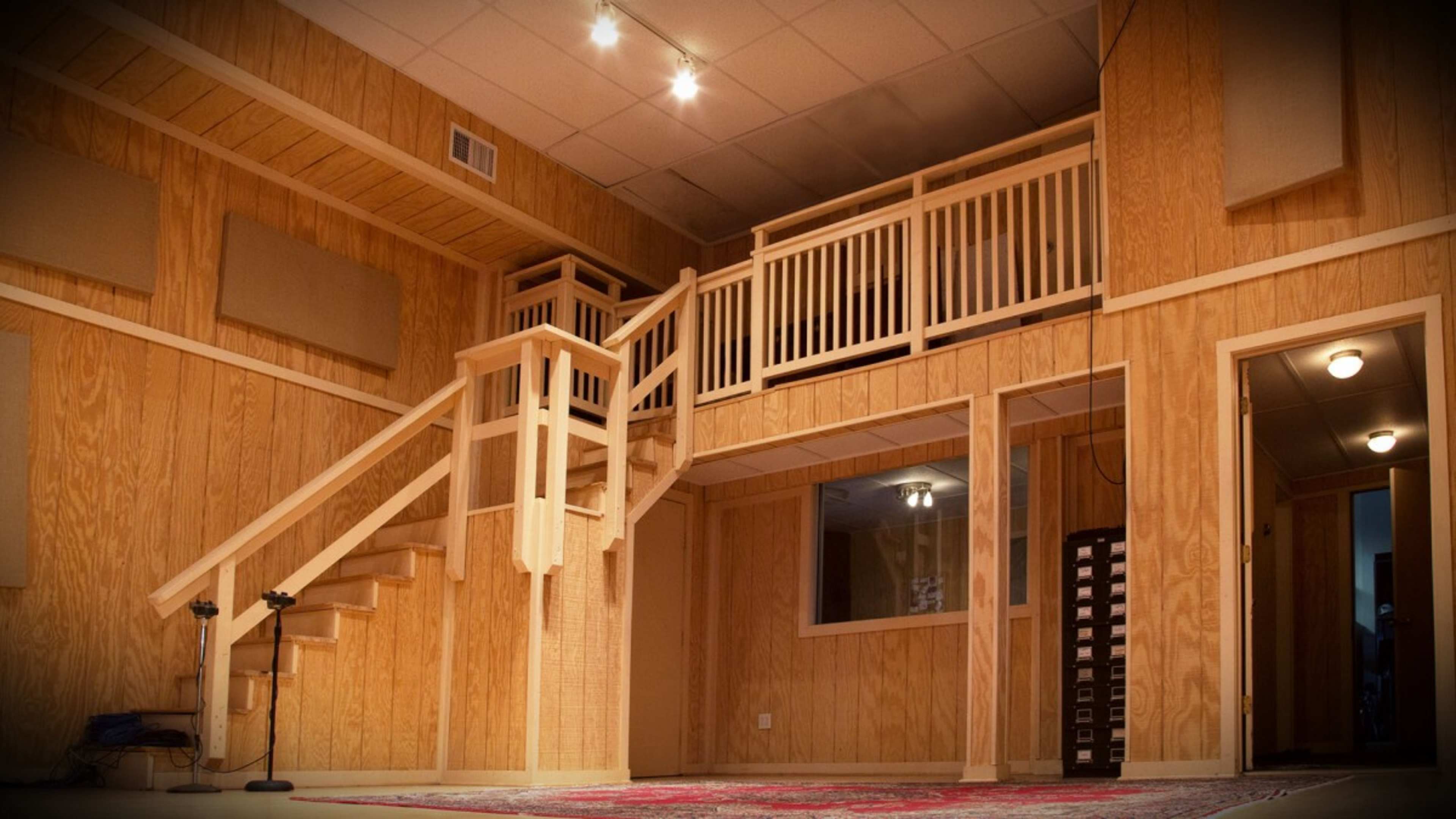 A wooden recording studio with a staircase leading to a balcony overlooking the main area, featuring soundproofing panels on the walls.