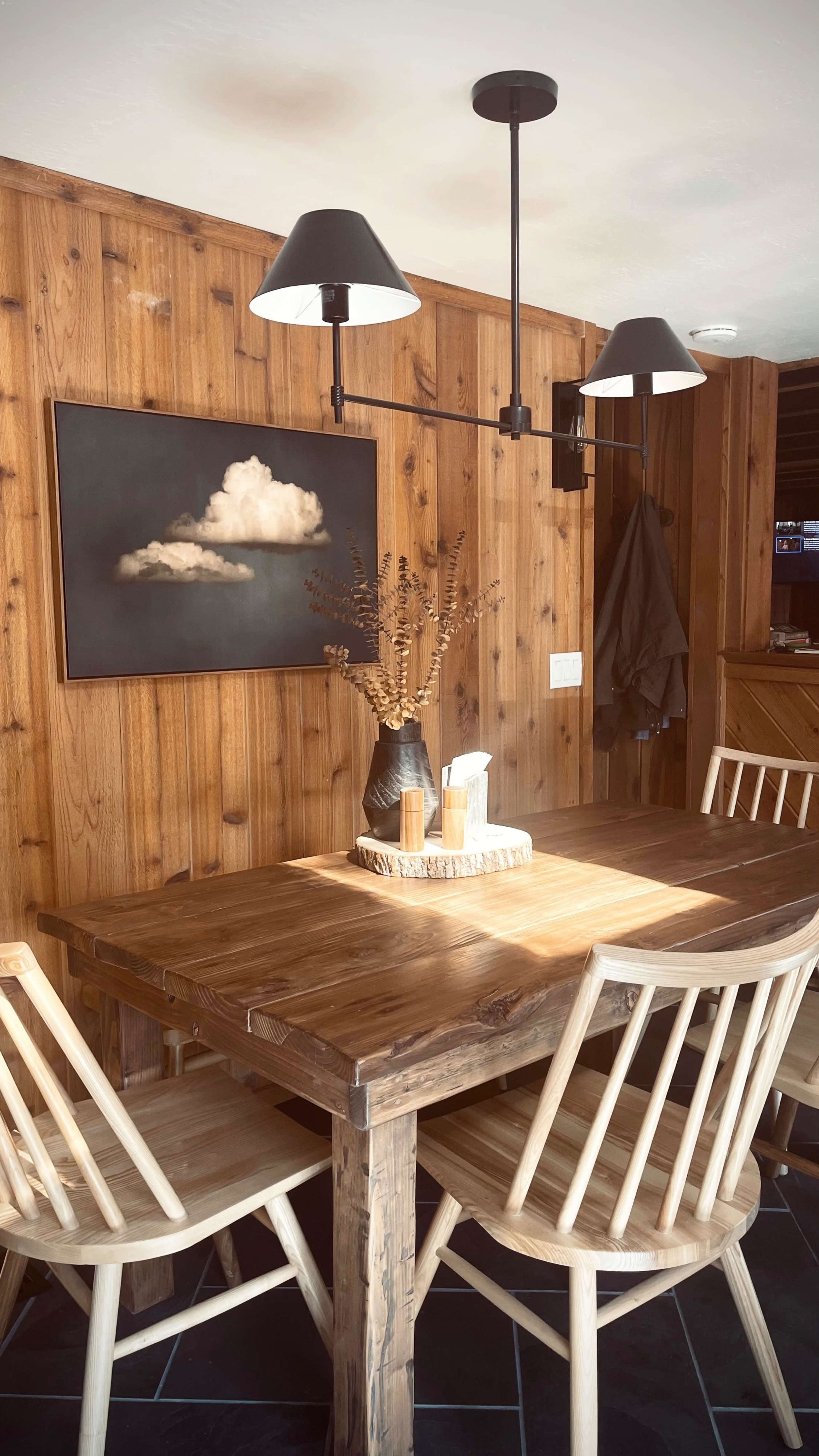 A wooden dining table surrounded by light-colored chairs is positioned against a wooden-paneled wall, with a painting of a cloud and a vase of dried flowers as decor.