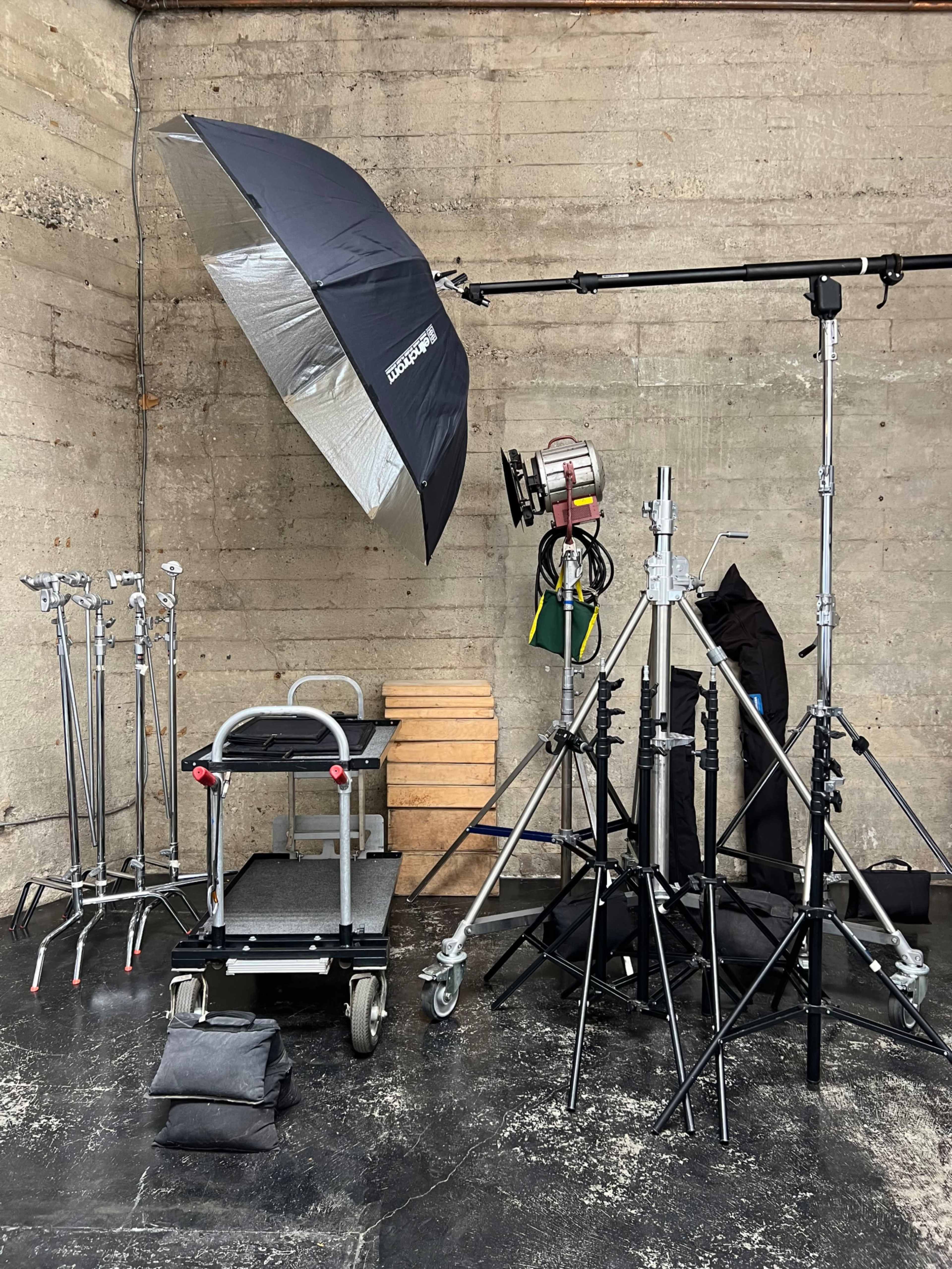 A photography studio setup features various light stands, a large umbrella reflector, and a cart surrounded by a concrete wall.