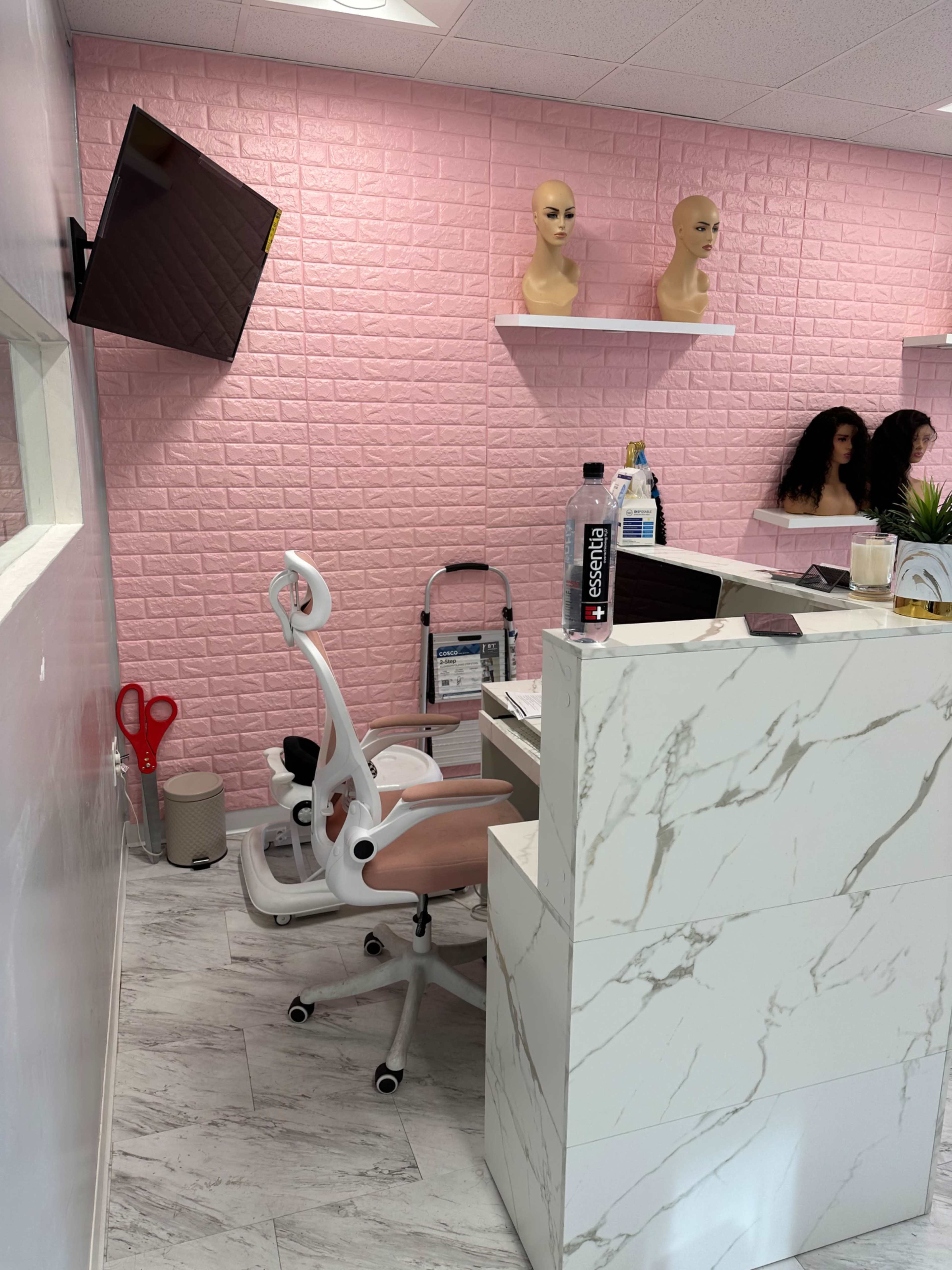 A salon interior with a pink brick wall, mannequins on shelves, and a modern white desk with a rolling chair.