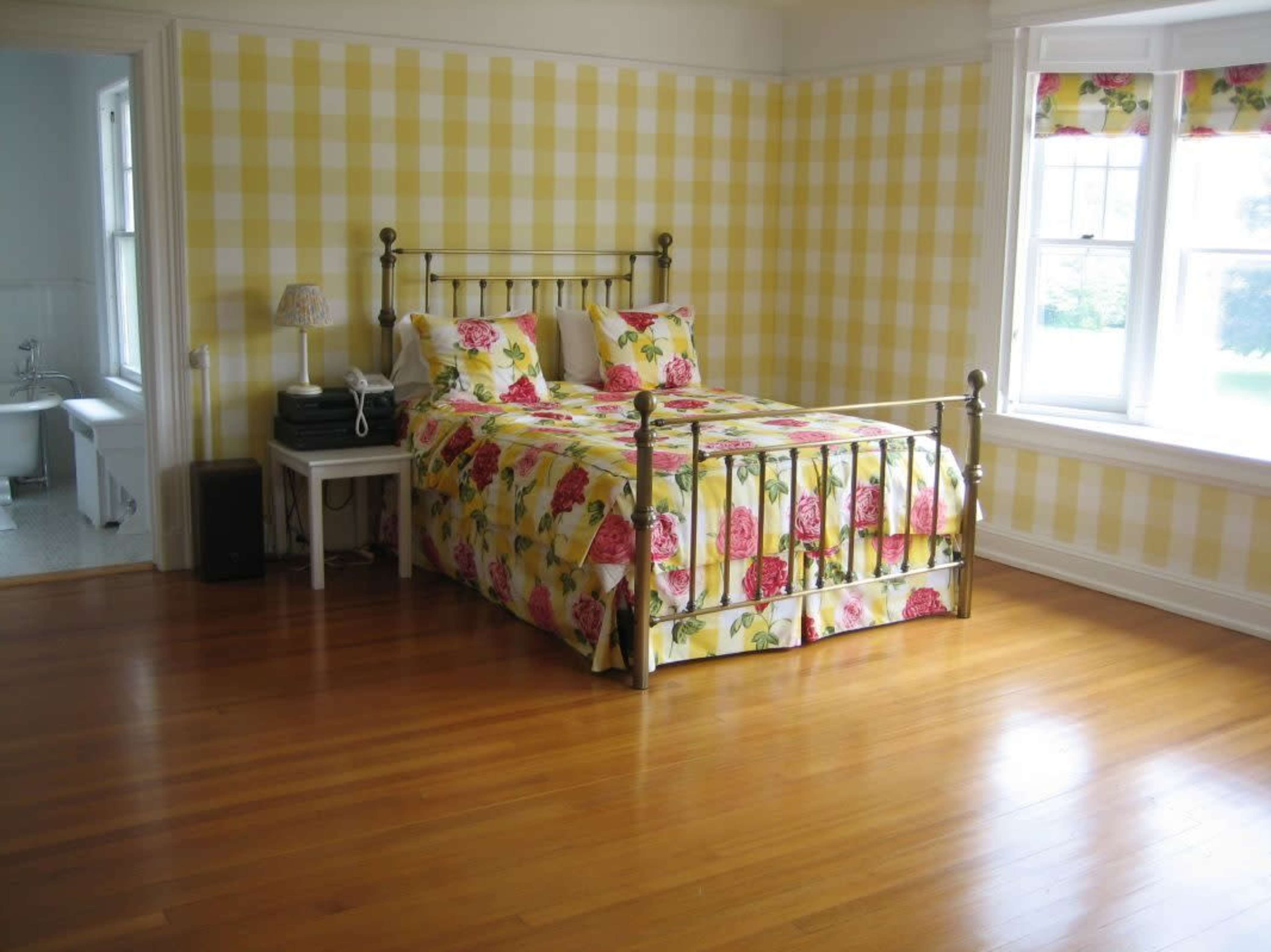 The image shows a bedroom with a bed covered in a floral pattern quilt, situated on wooden flooring, against a yellow checkered wall.