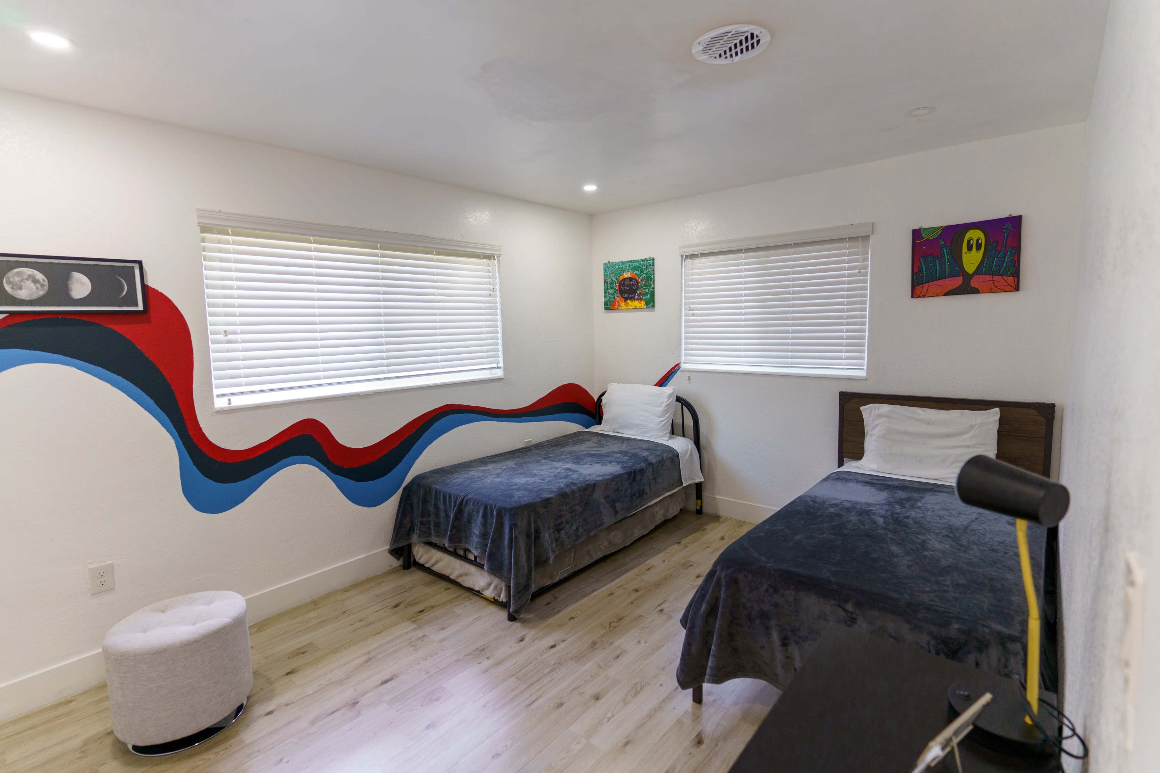 A minimalist bedroom with two twin beds, a small round ottoman, and colorful wall art, illuminated by natural light through the window.
