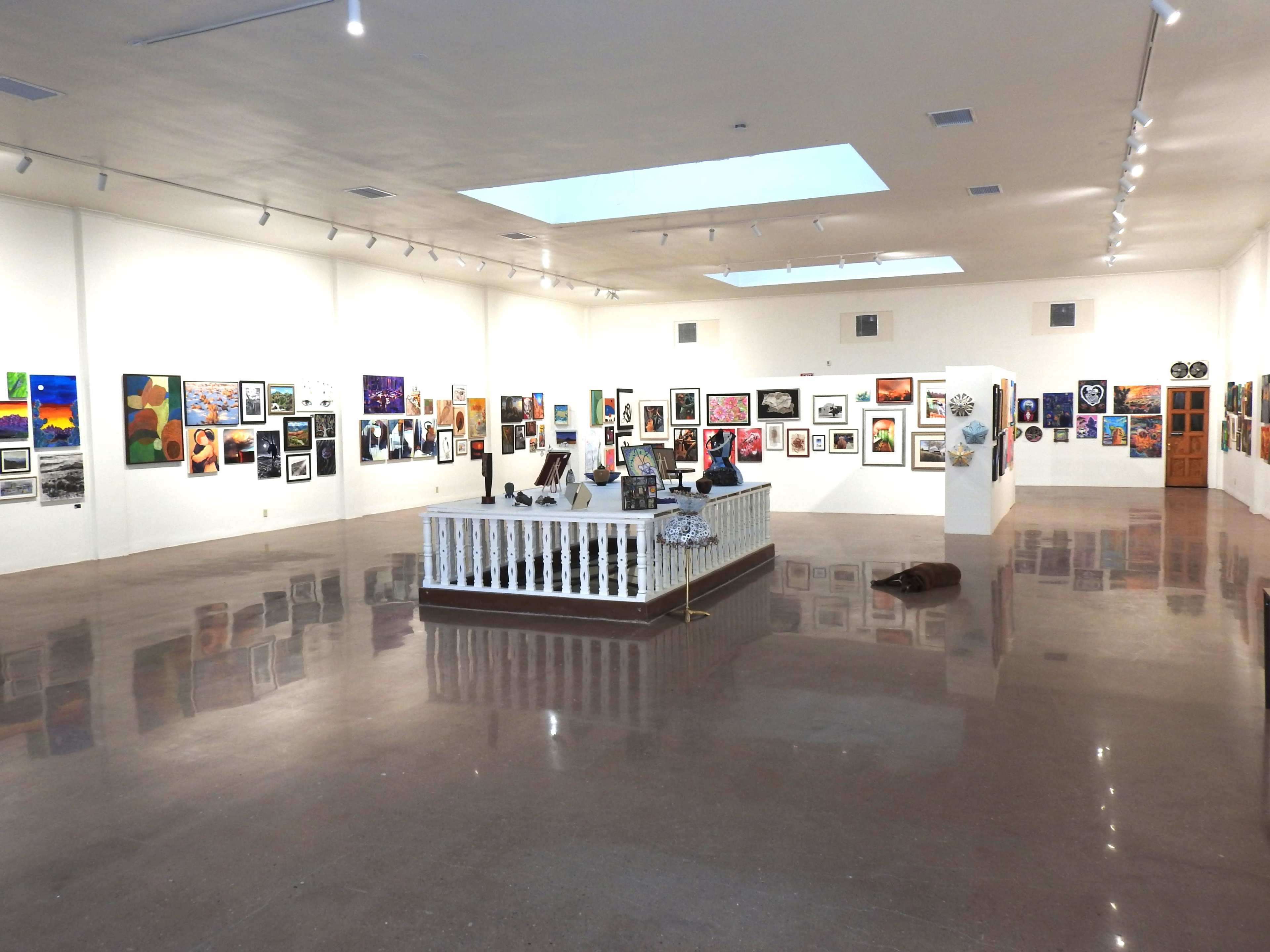The image shows an art gallery filled with various framed artworks displayed on white walls, with a central white fence enclosing a small area.