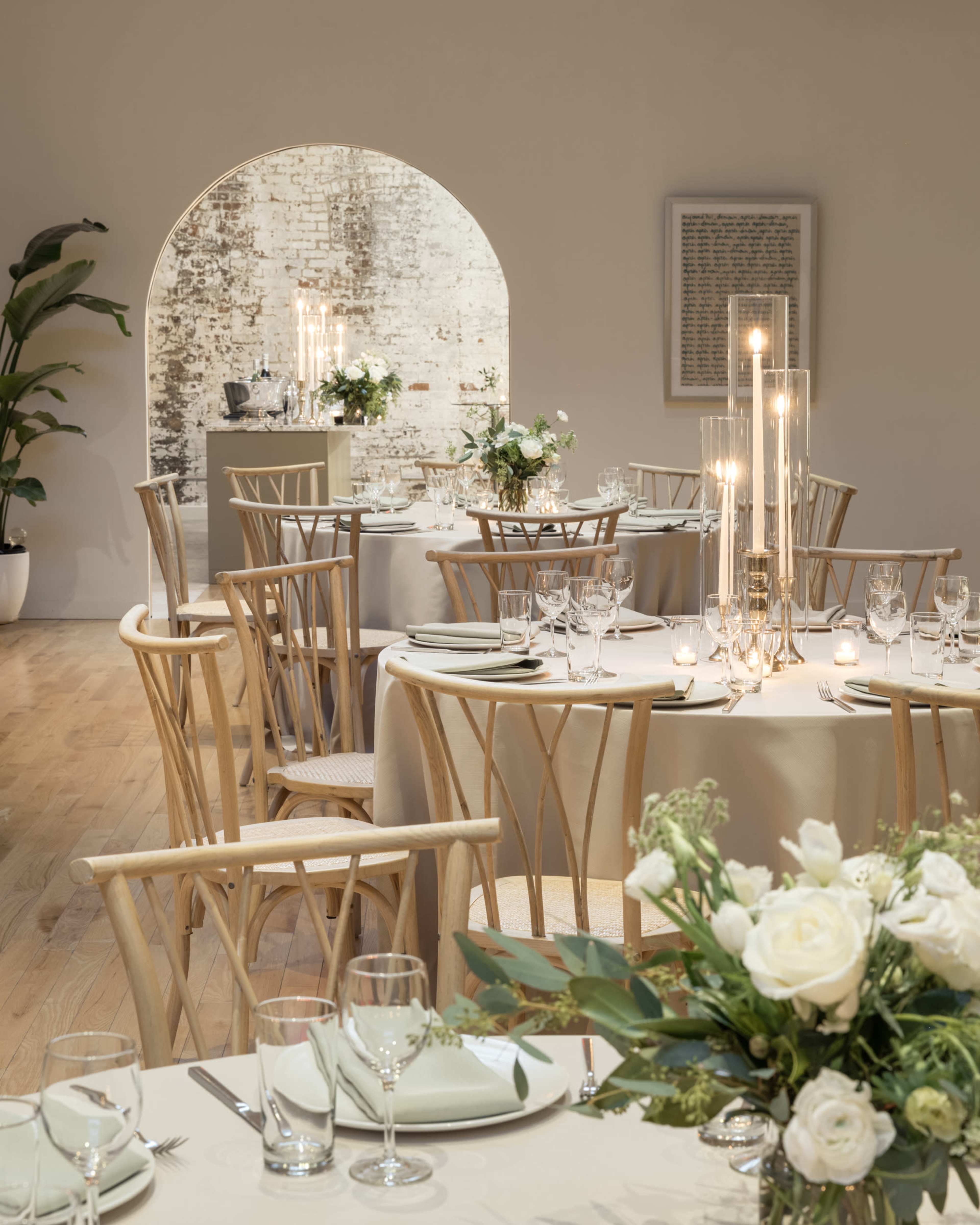 A dining area with elegantly set tables, featuring wooden chairs and floral centerpieces, alongside an arched entrance revealing a decorated wall and additional furnishings.