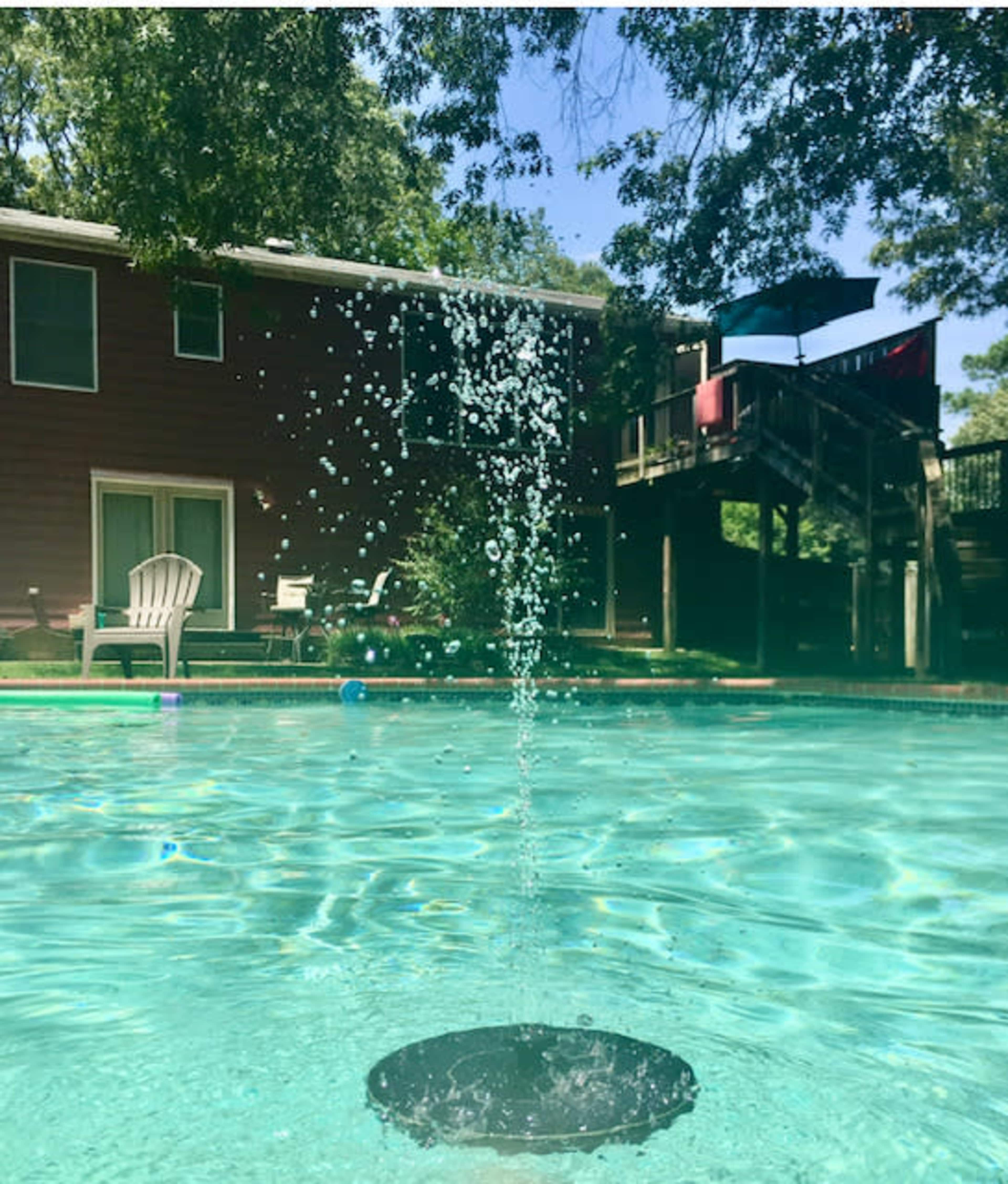A fountain sprays water into a swimming pool, with a backyard and house visible in the background.