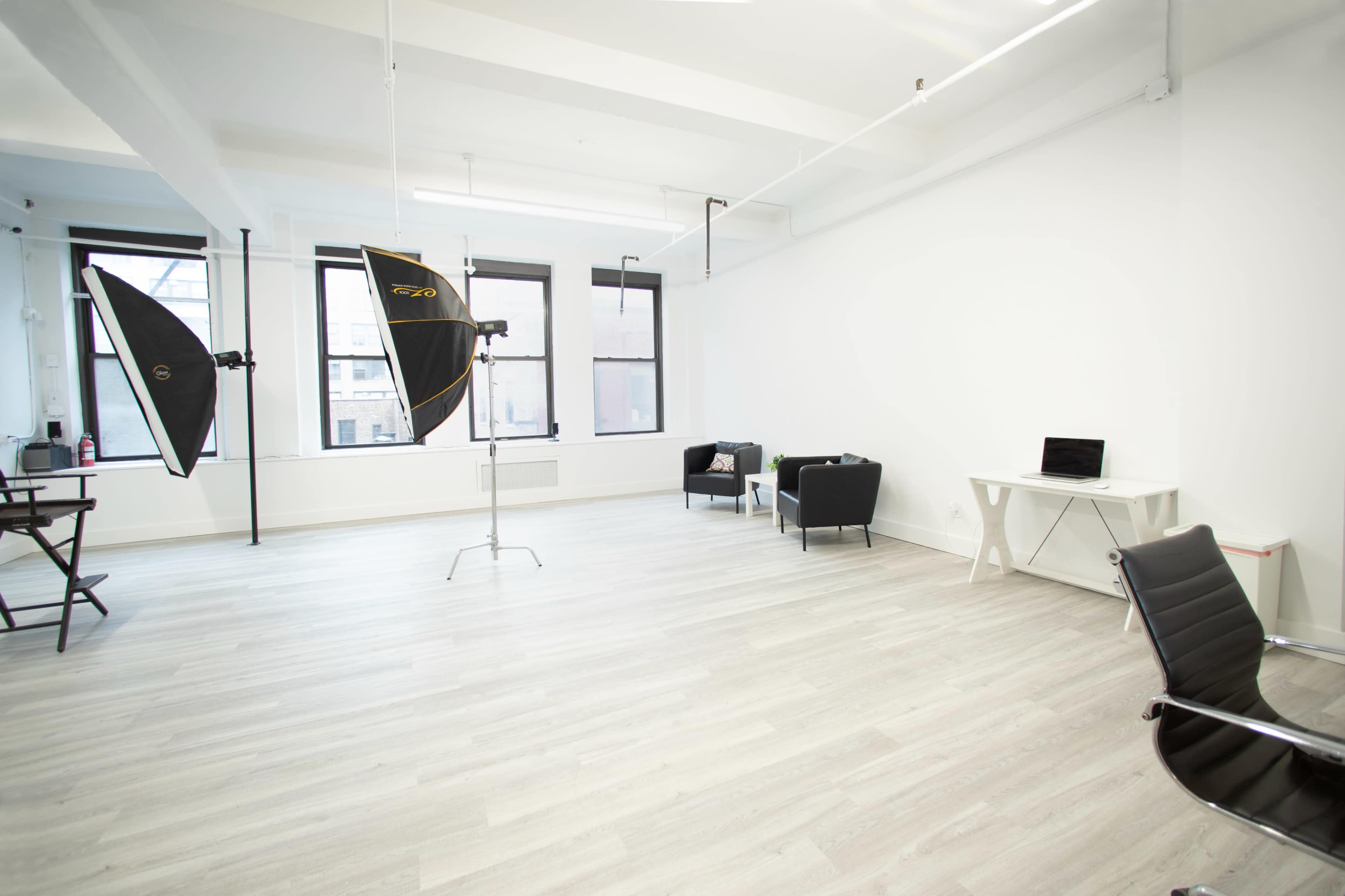 A spacious, well-lit studio with light gray flooring, two softbox lights on stands, a black chair, a black sofa, a white desk with a laptop, and large windows.