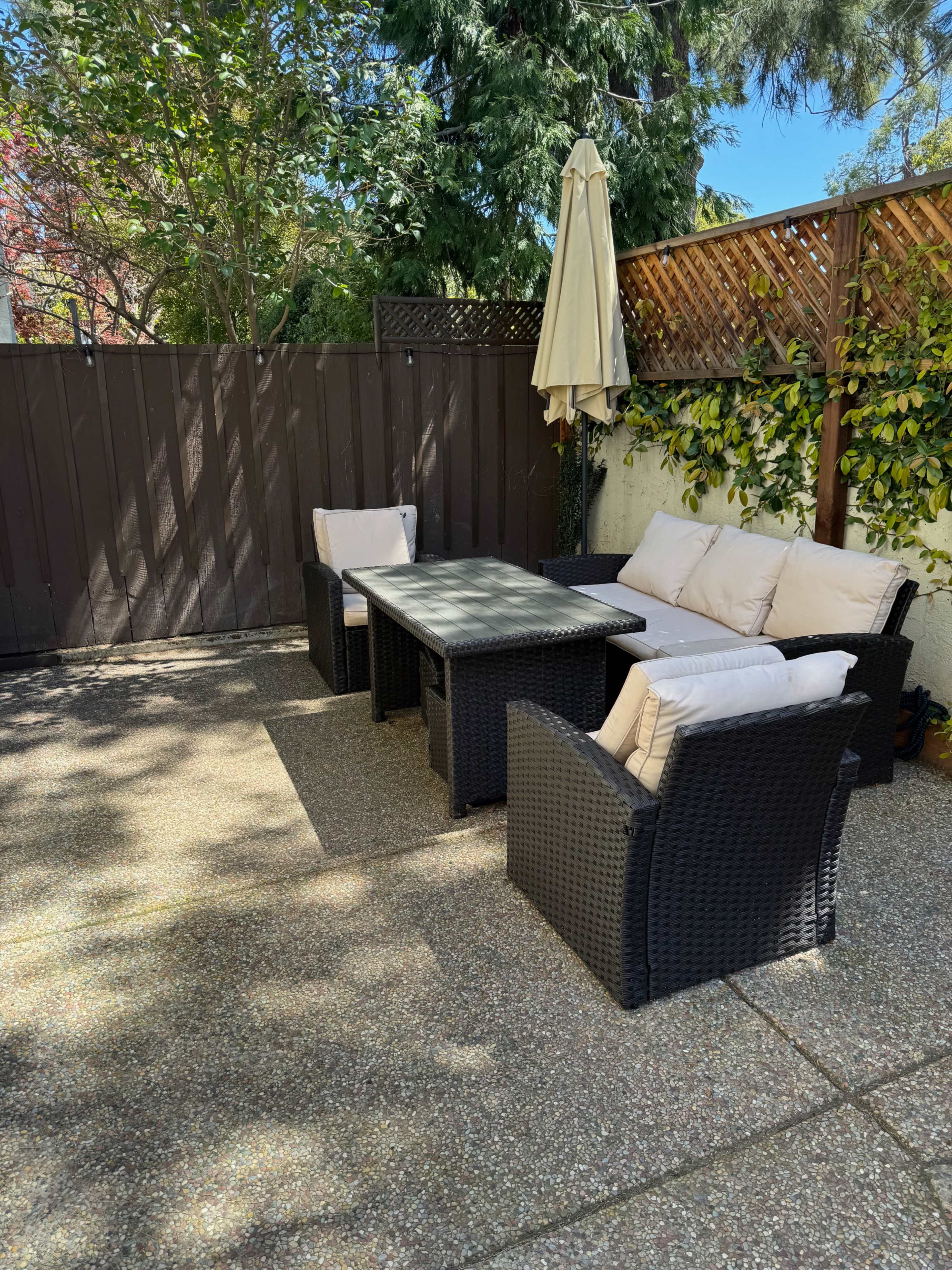 A patio area features a rectangular table surrounded by four wicker chairs and a beige umbrella, enclosed by a wooden fence and greenery.