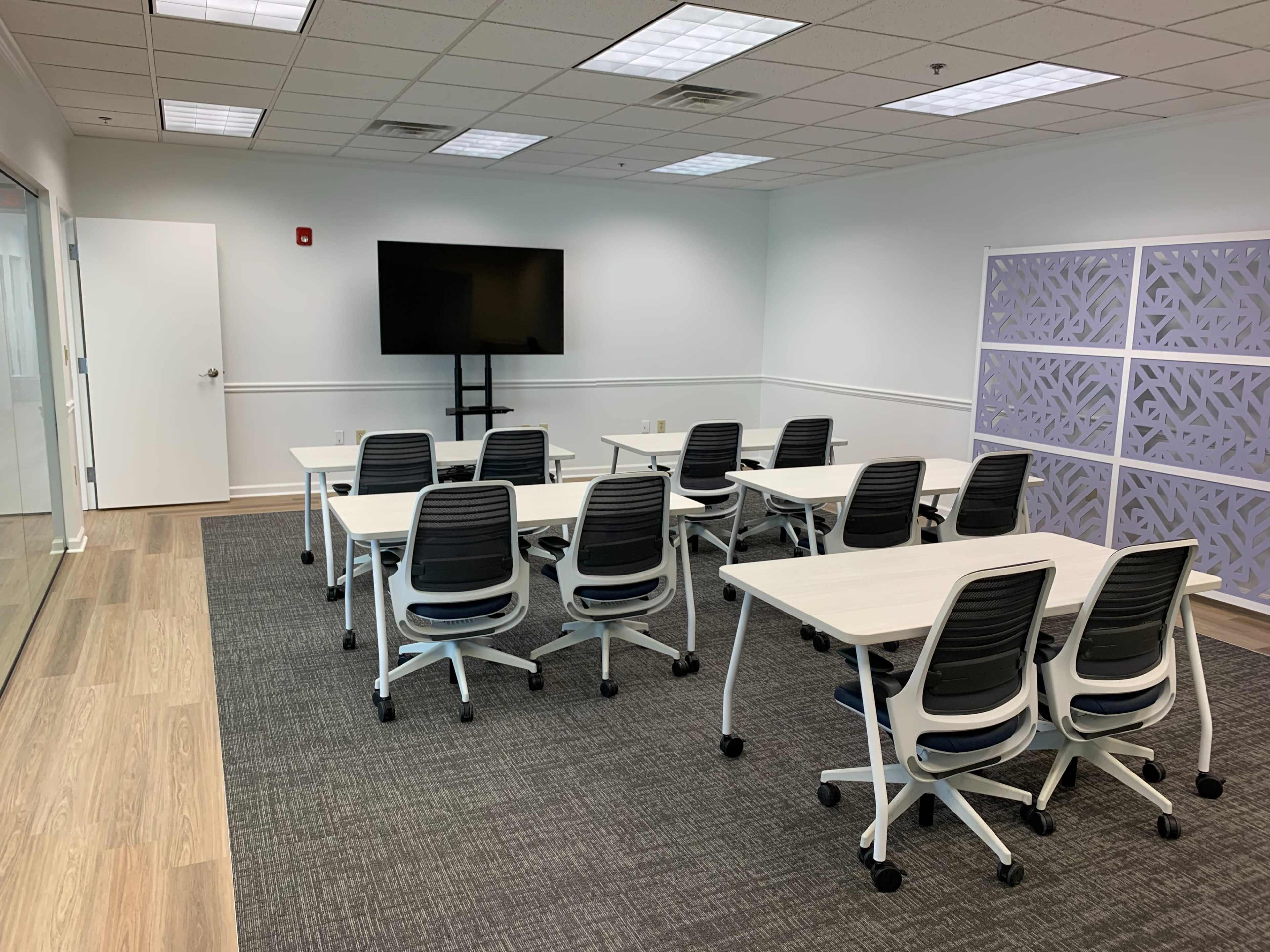 A bright, modern meeting room with several tables and chairs arranged facing a wall-mounted television.