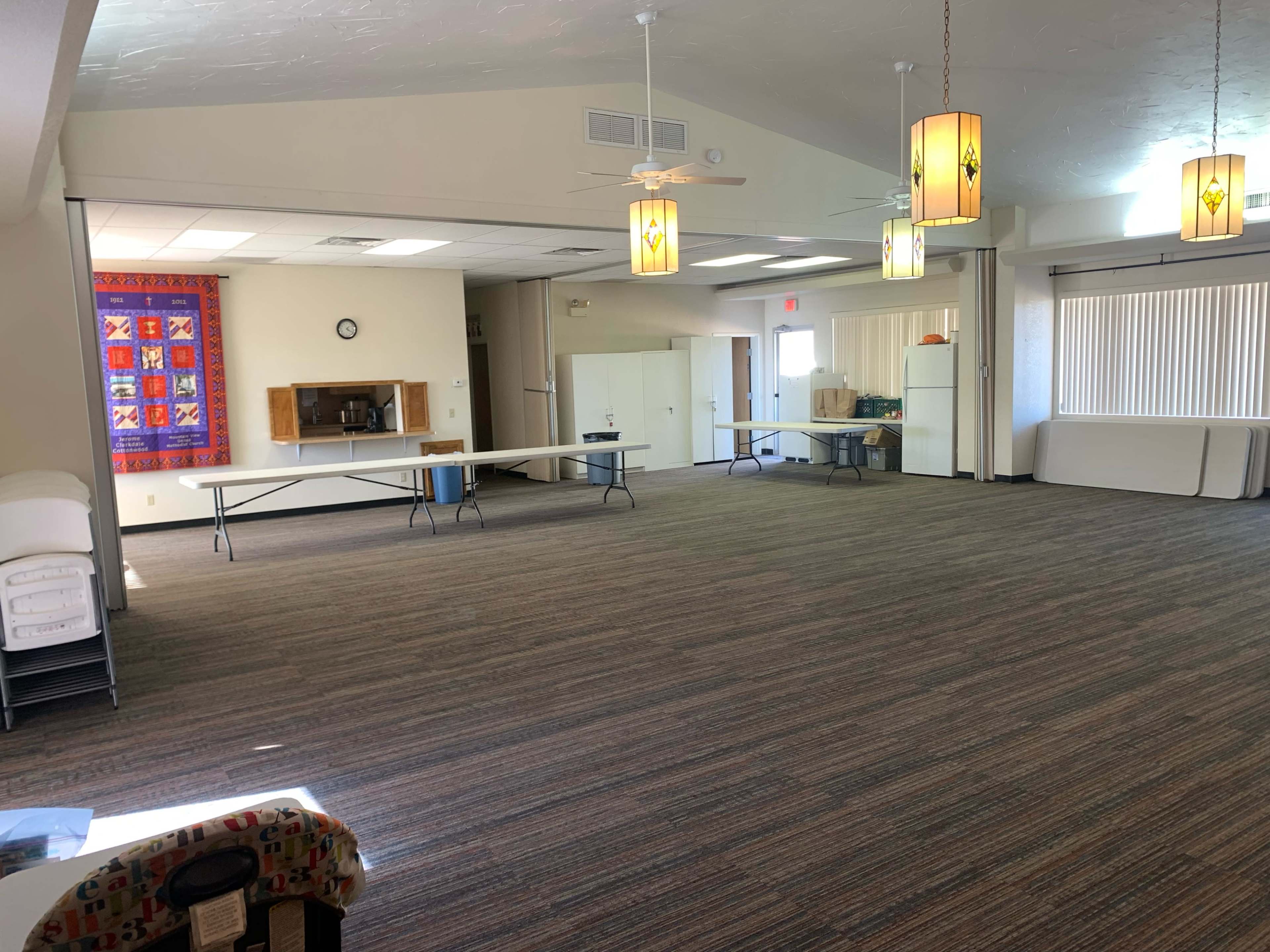 A spacious multipurpose room with tables arranged for use, a kitchenette area, and decorative lighting.