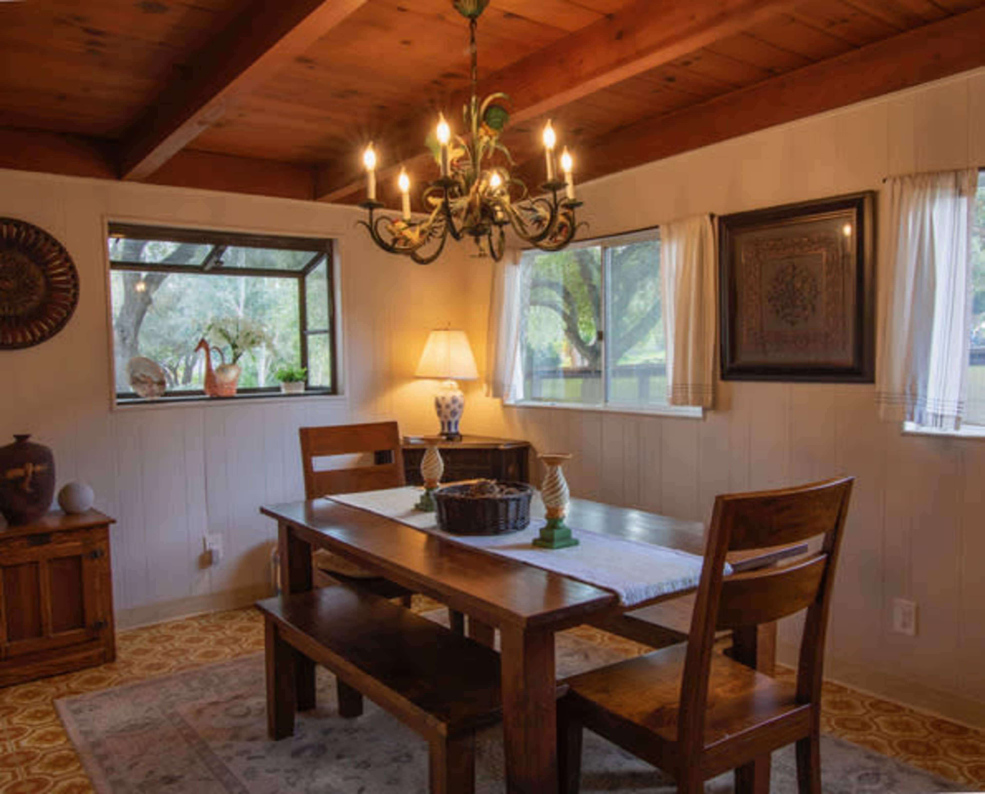 The image shows a cozy dining area featuring a wooden table with benches, a chandelier, and windows overlooking greenery.