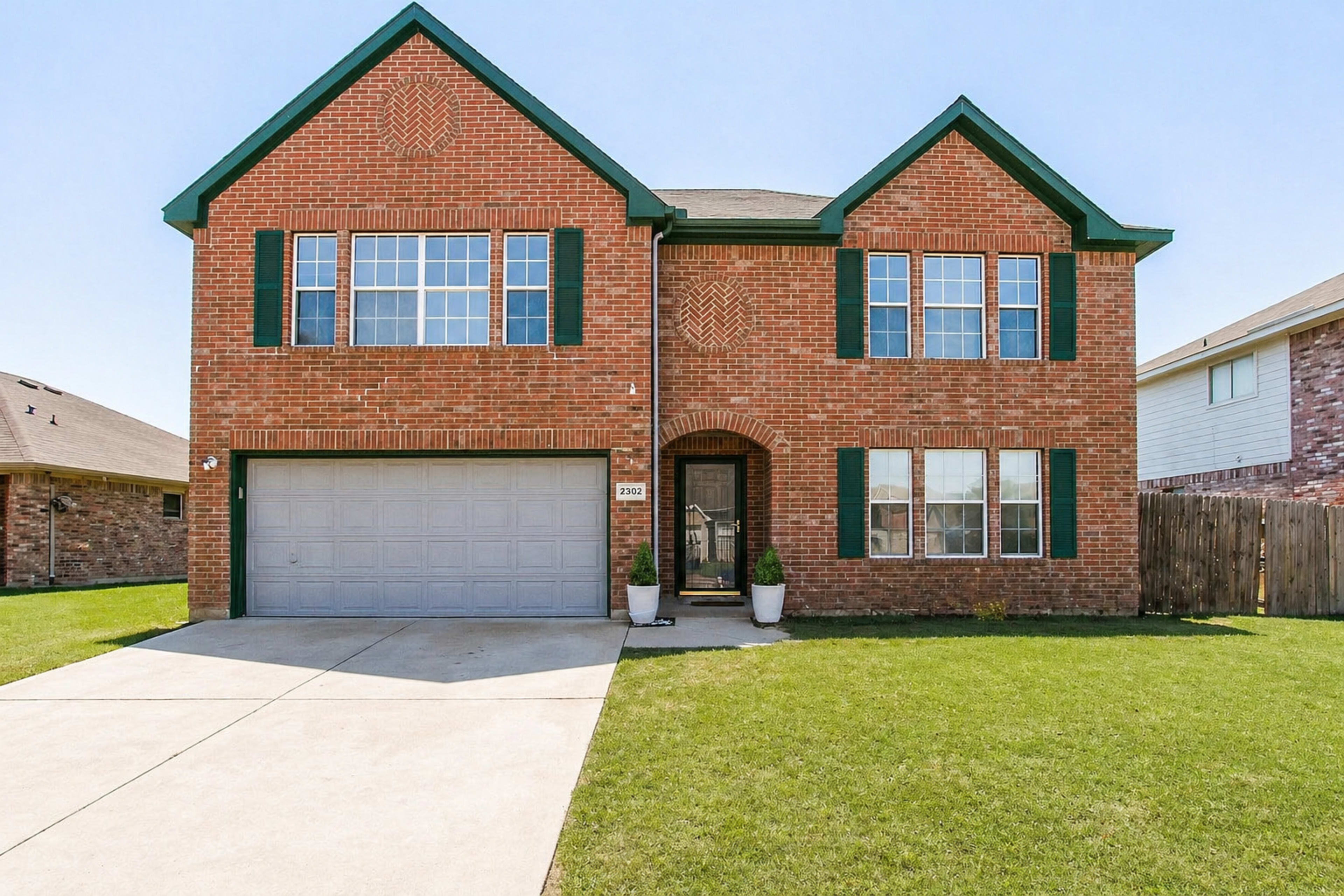 The image shows a two-story brick house with green shutters, a double garage, and a well-maintained lawn.