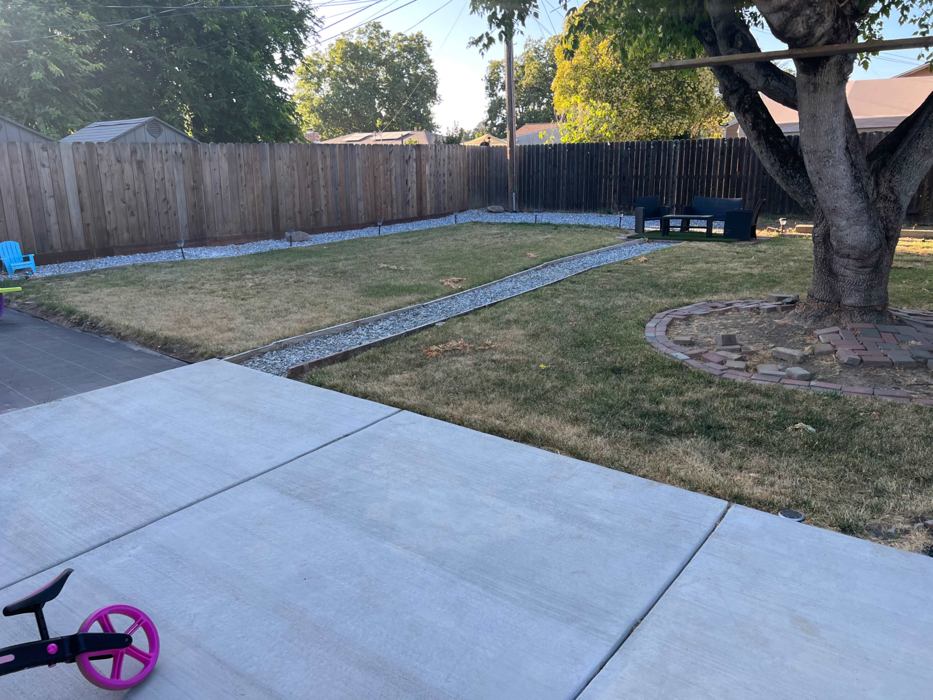 A grassy backyard features a gravel path leading to a seating area, surrounded by wooden fencing and a tree.