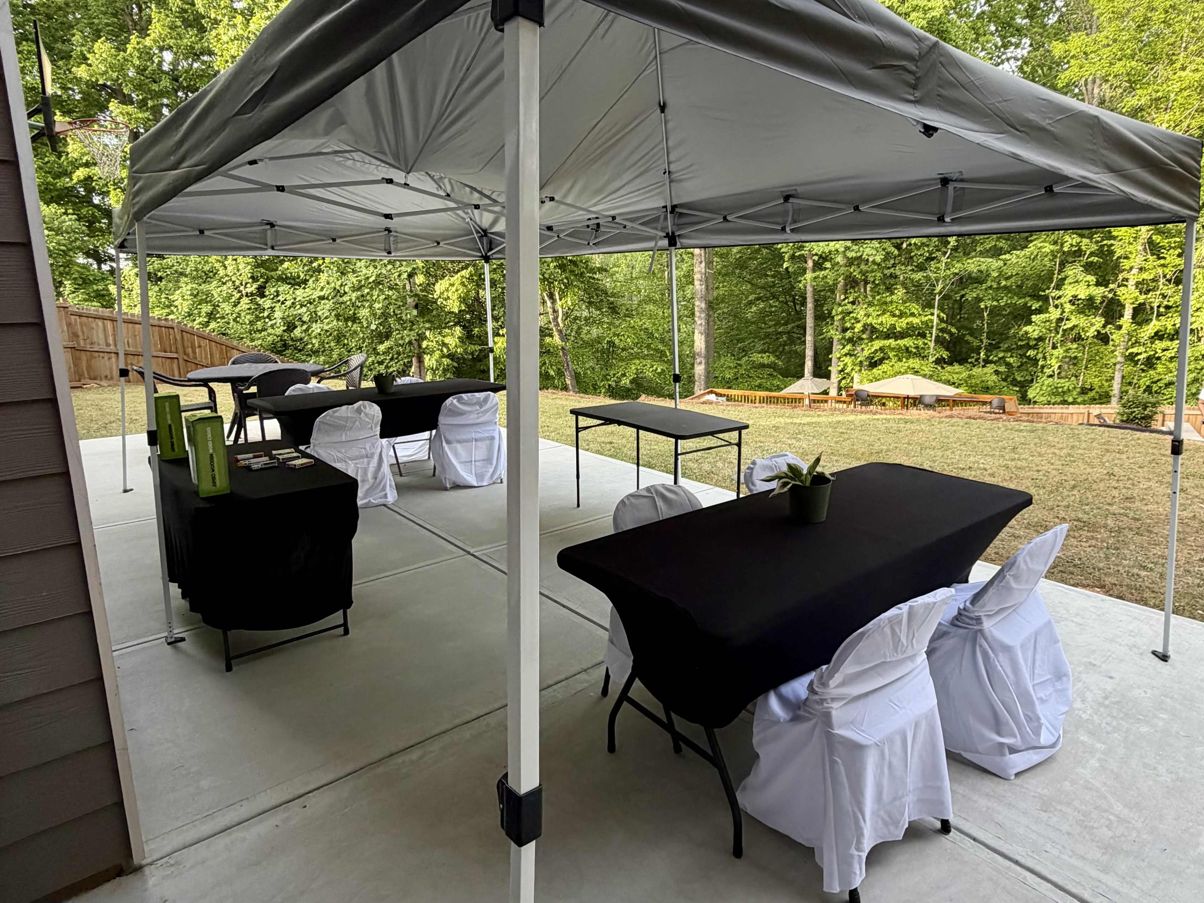 The image shows a covered outdoor area with several black and white tables and chairs arranged, surrounded by green trees.