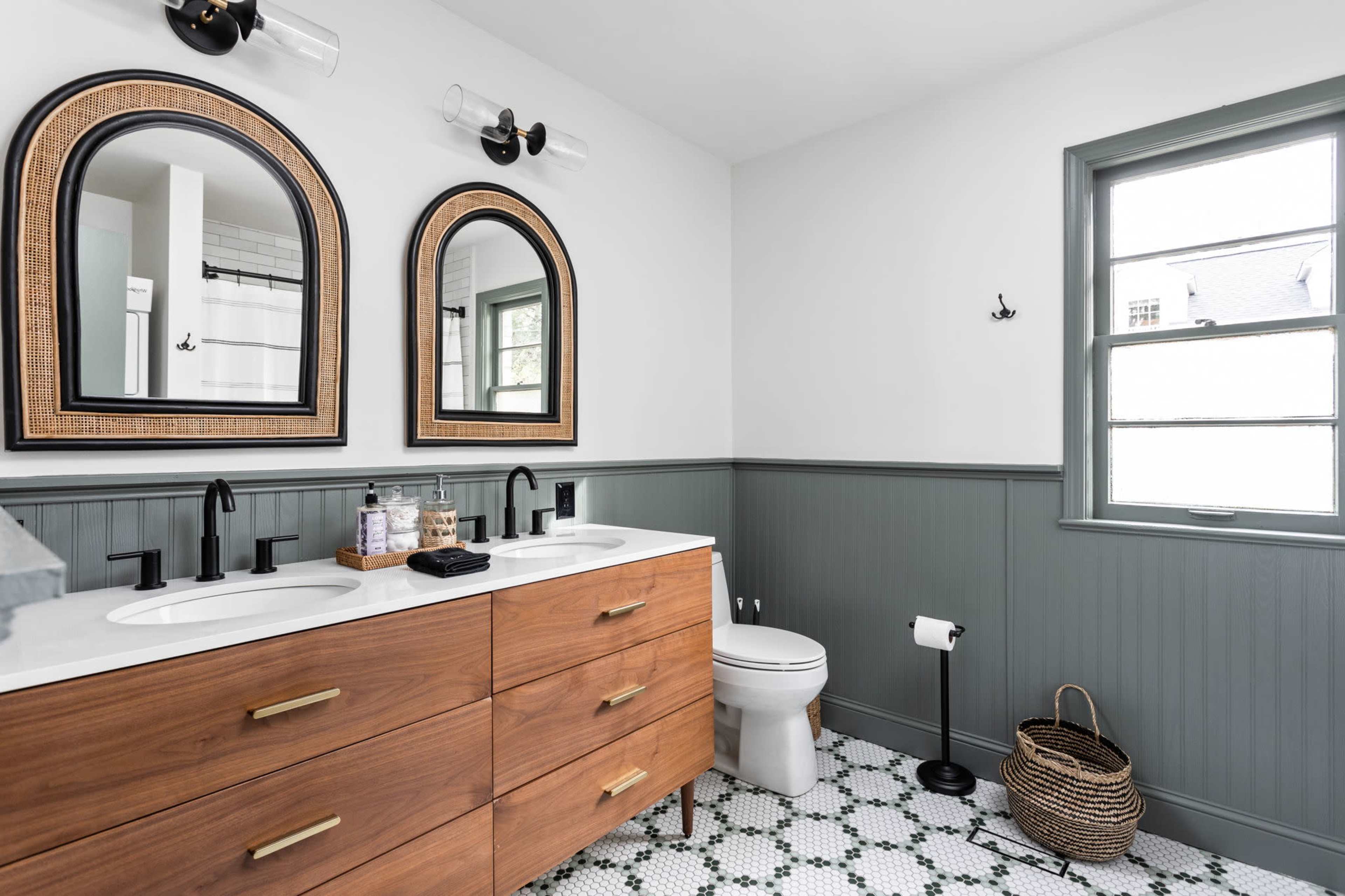 A modern bathroom featuring double sinks in a wooden vanity beneath two framed mirrors, with a patterned floor, a window, and a toilet.