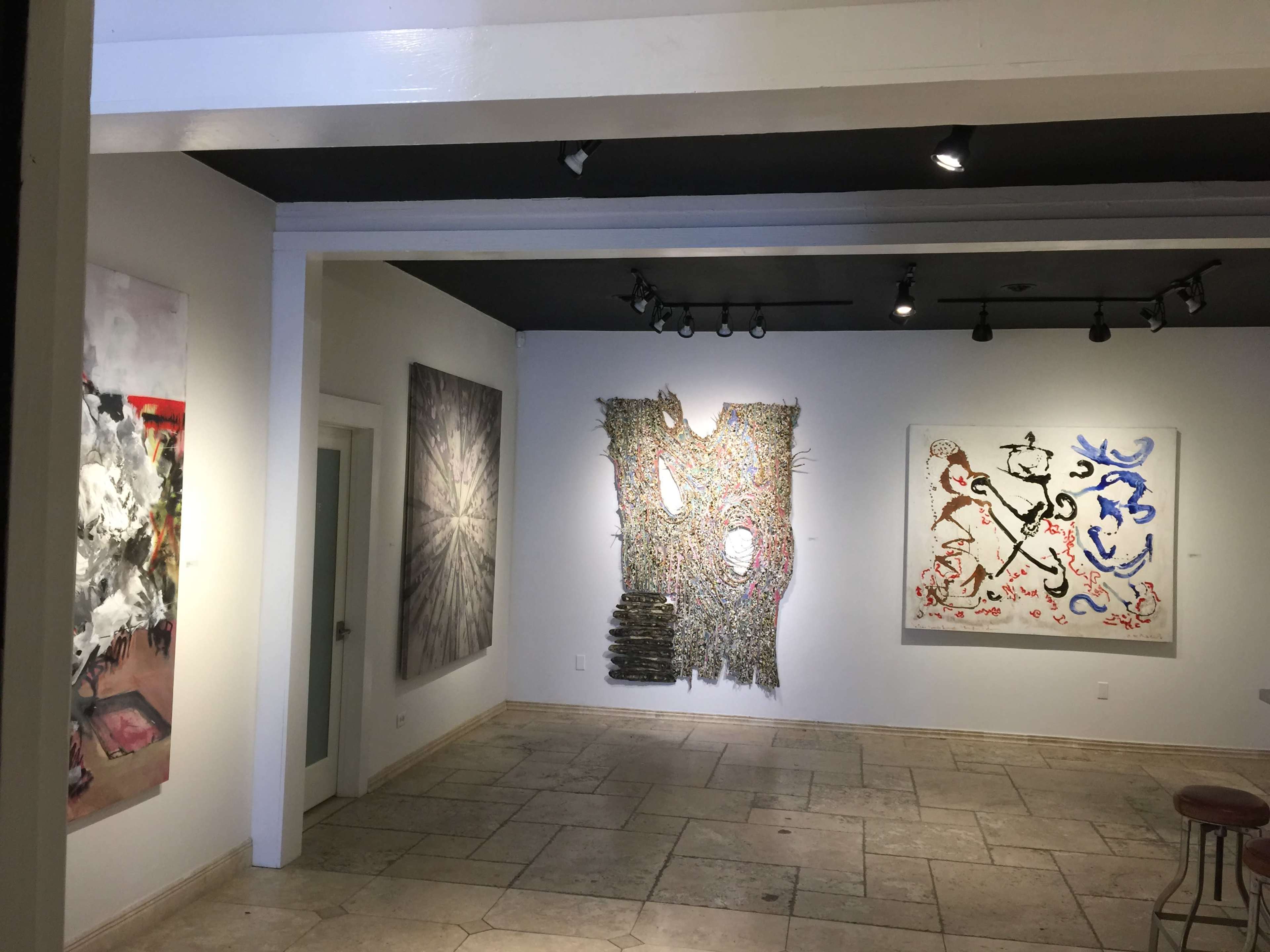 The image shows an art gallery with several colorful artworks displayed on the walls, including paintings and mixed media pieces, and a stone floor.