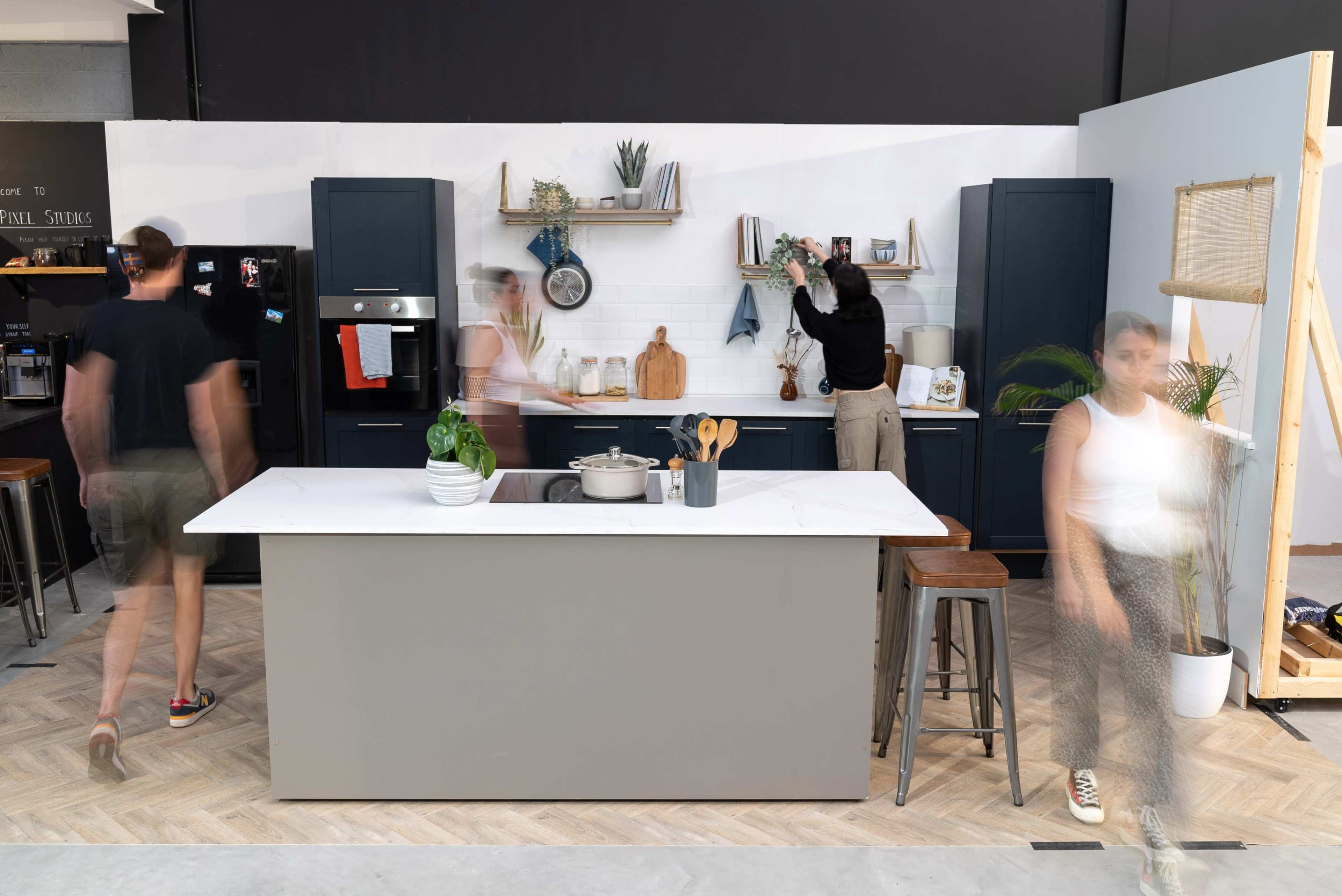 A modern kitchen featuring an island, dark cabinetry, and individuals engaged in various activities, with a lively atmosphere.