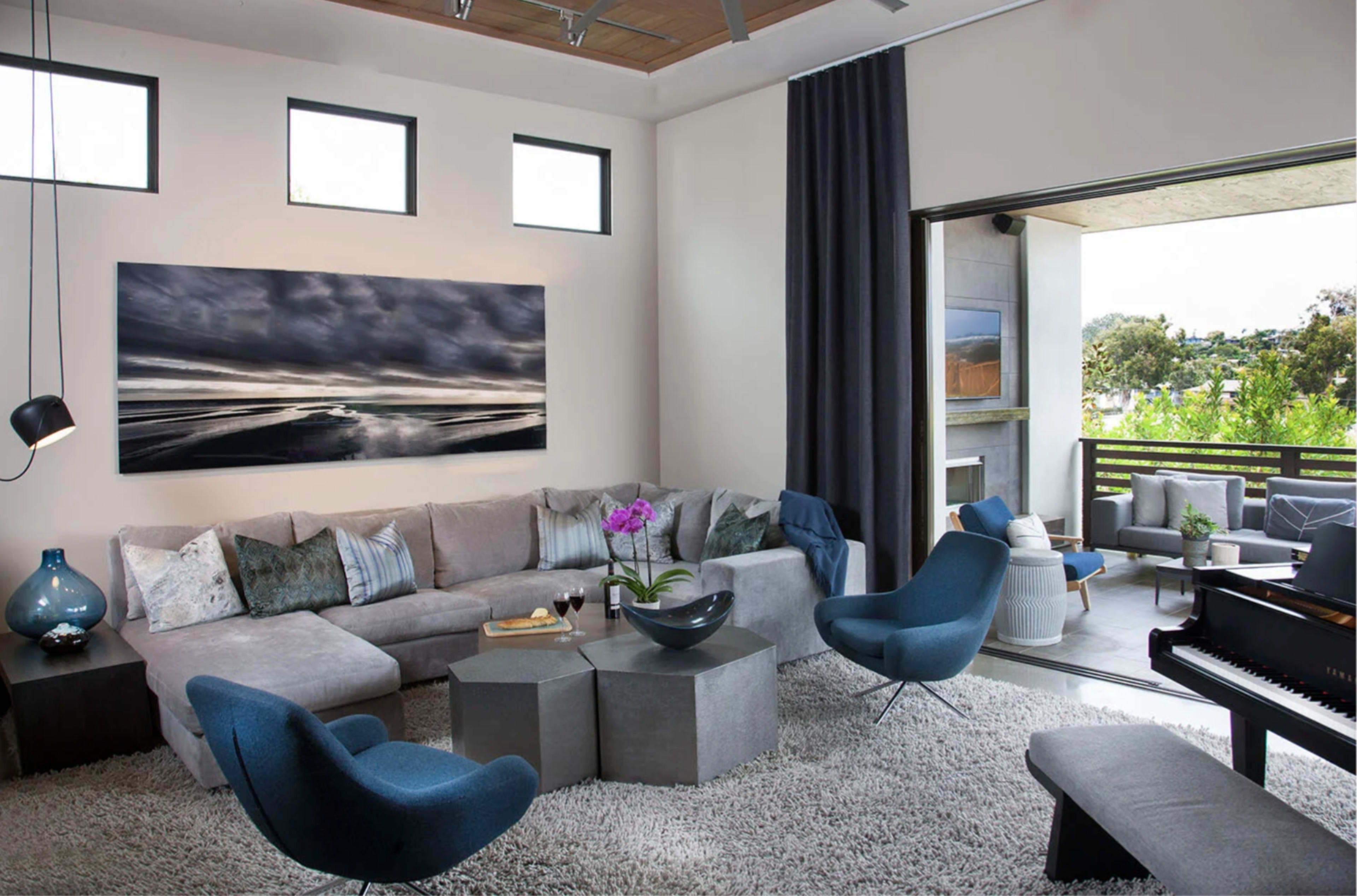 A modern living room features a sectional sofa, circular coffee tables, and a piano, with large windows providing natural light and views of a patio.