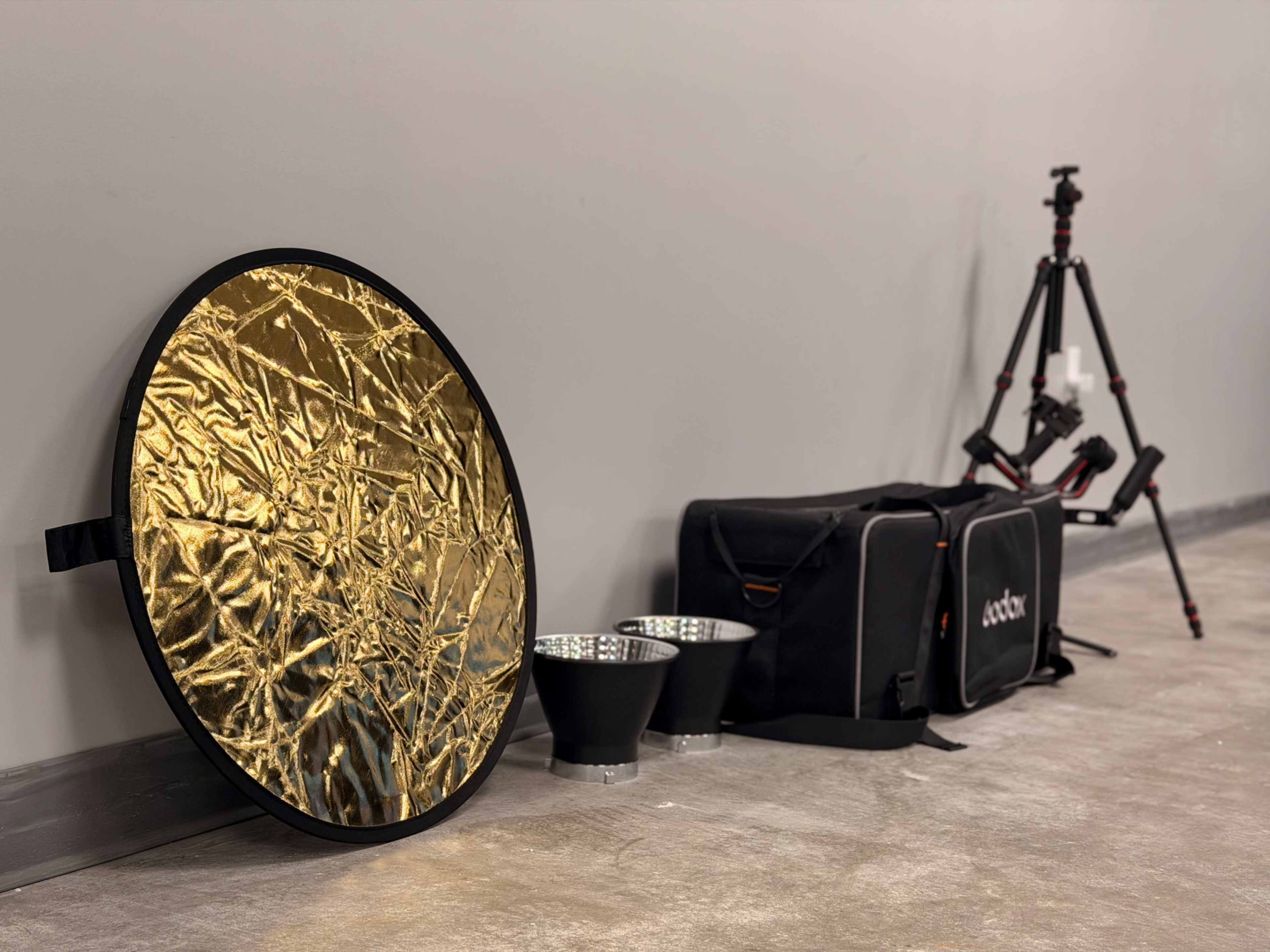 The image shows a photography setup that includes a gold reflector, two light stands, a camera bag, and a tripod resting against a gray wall.