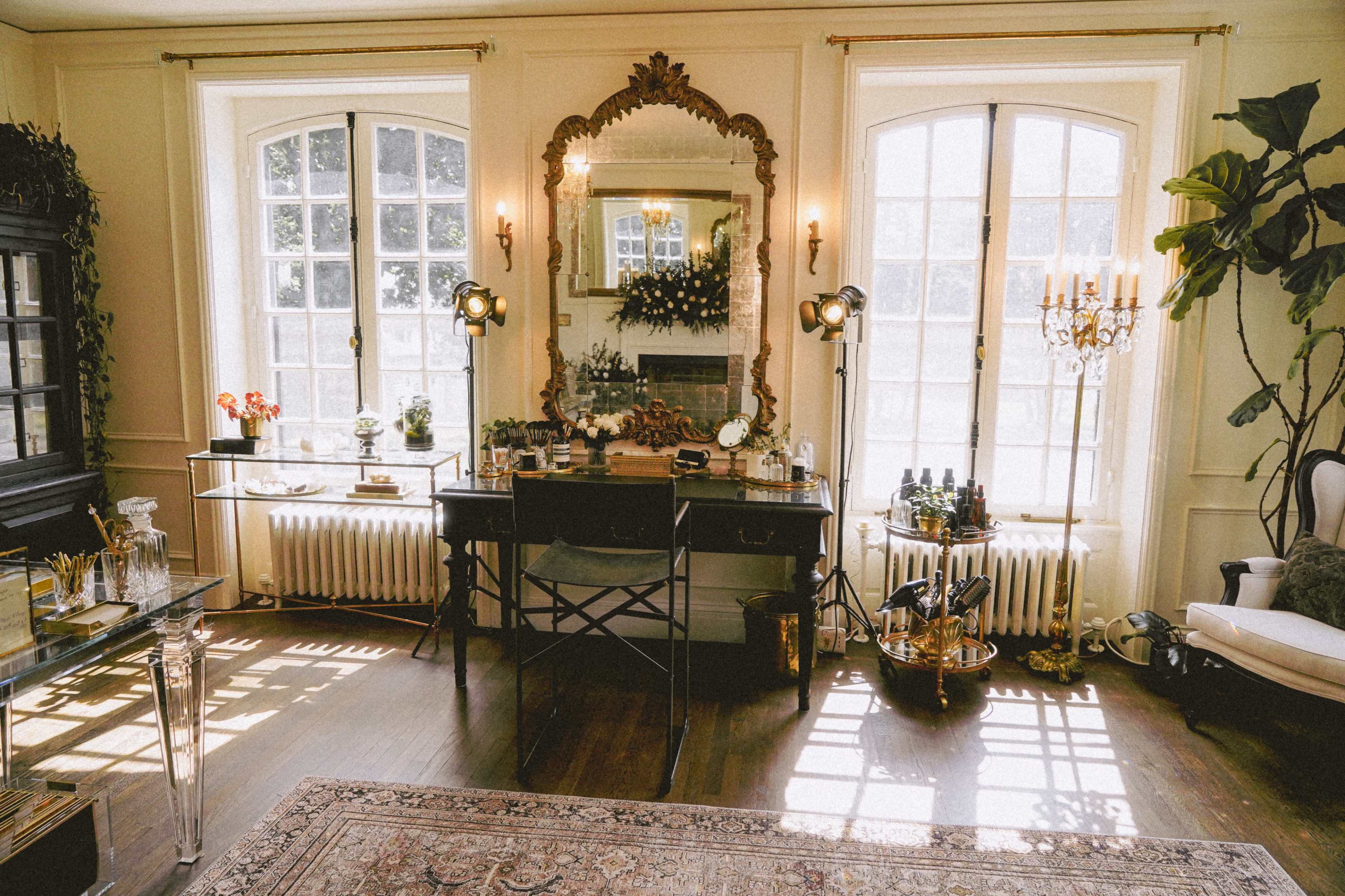 A well-lit room features a vintage vanity table with a large ornate mirror, surrounded by decorative plants and various beauty products.