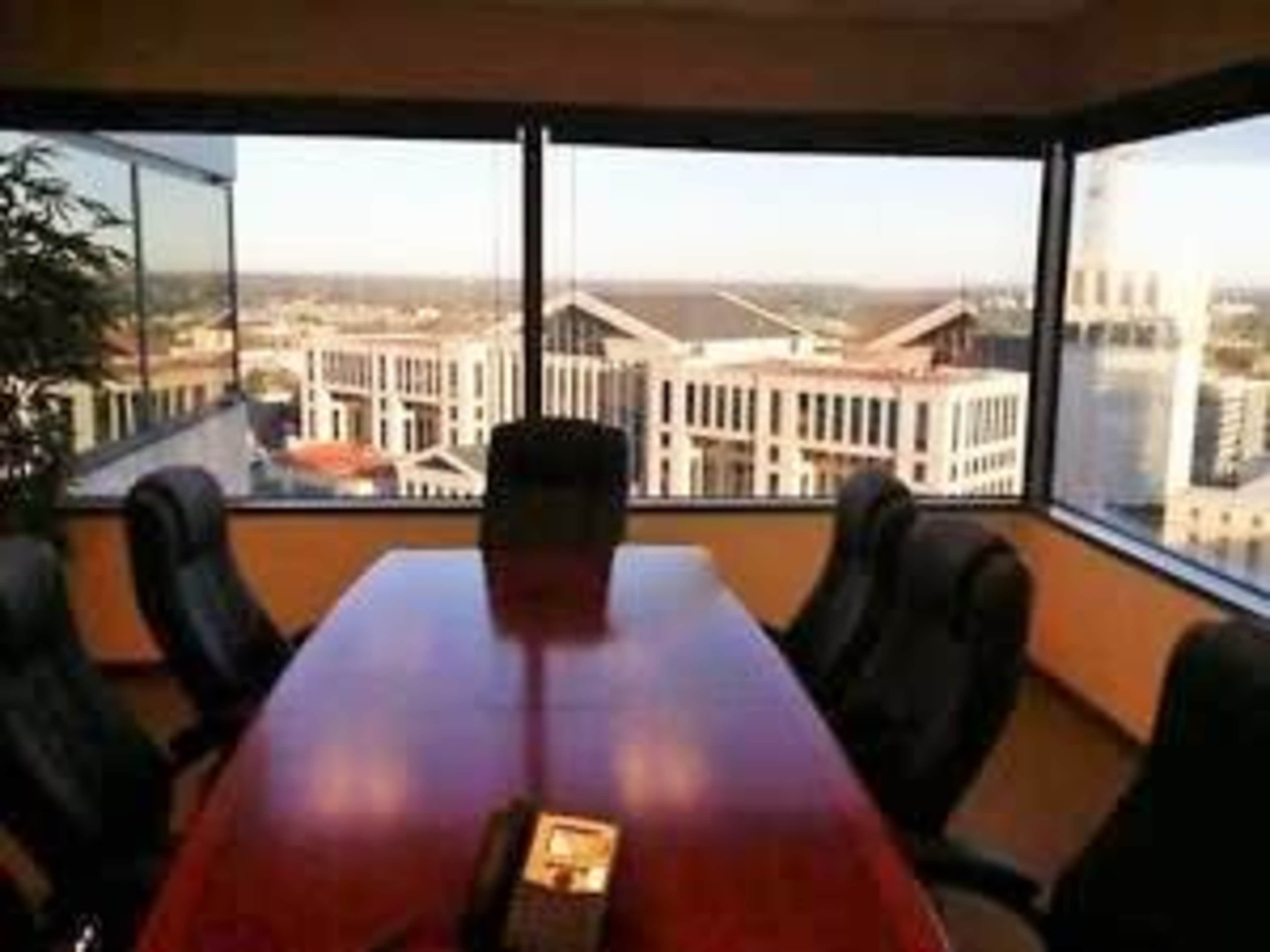The image shows a conference room with a large wooden table surrounded by black leather chairs, featuring large windows that provide a view of a cityscape.
