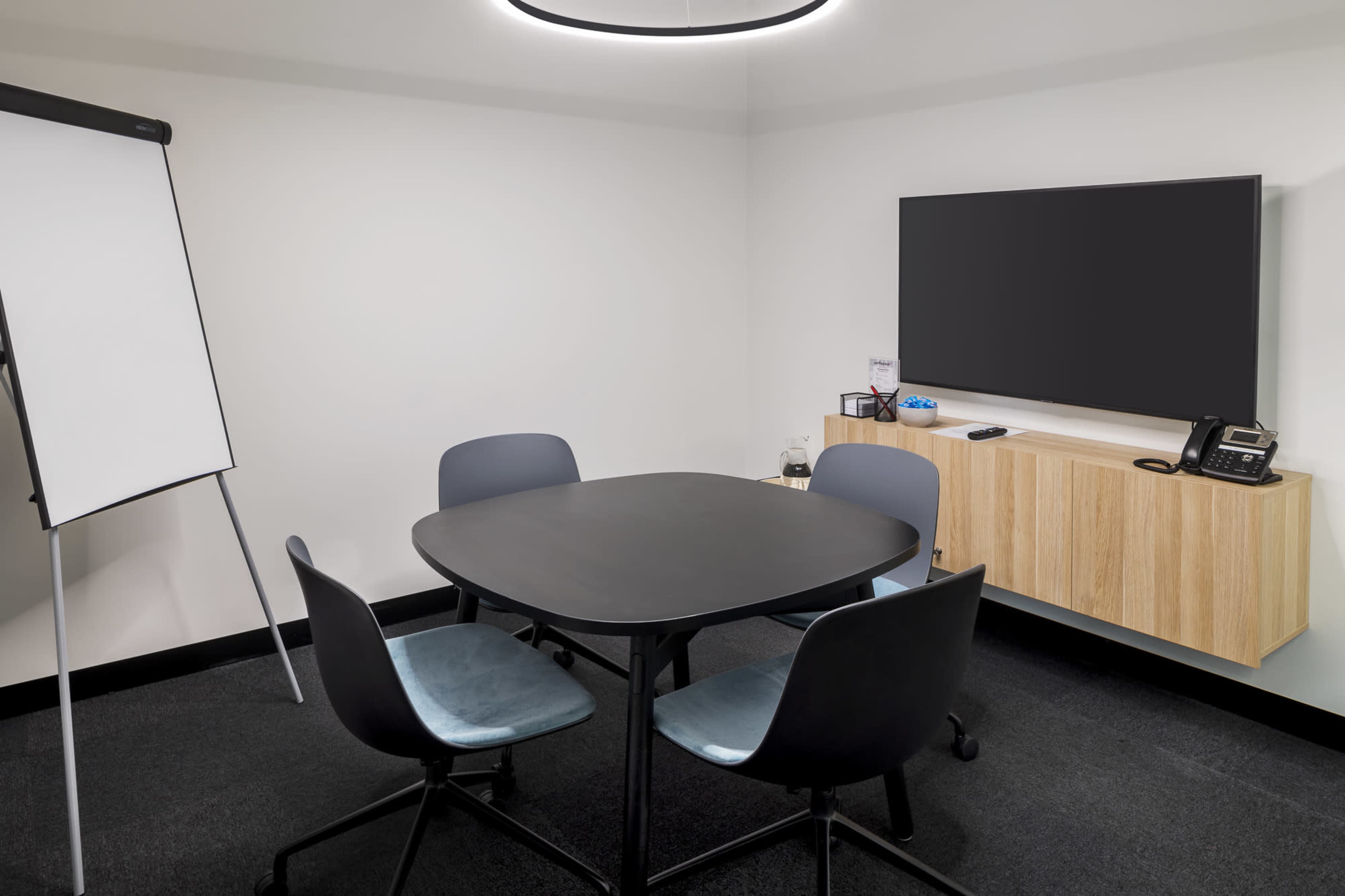A small conference room features a round black table surrounded by four chairs, a whiteboard, and a wall-mounted TV.