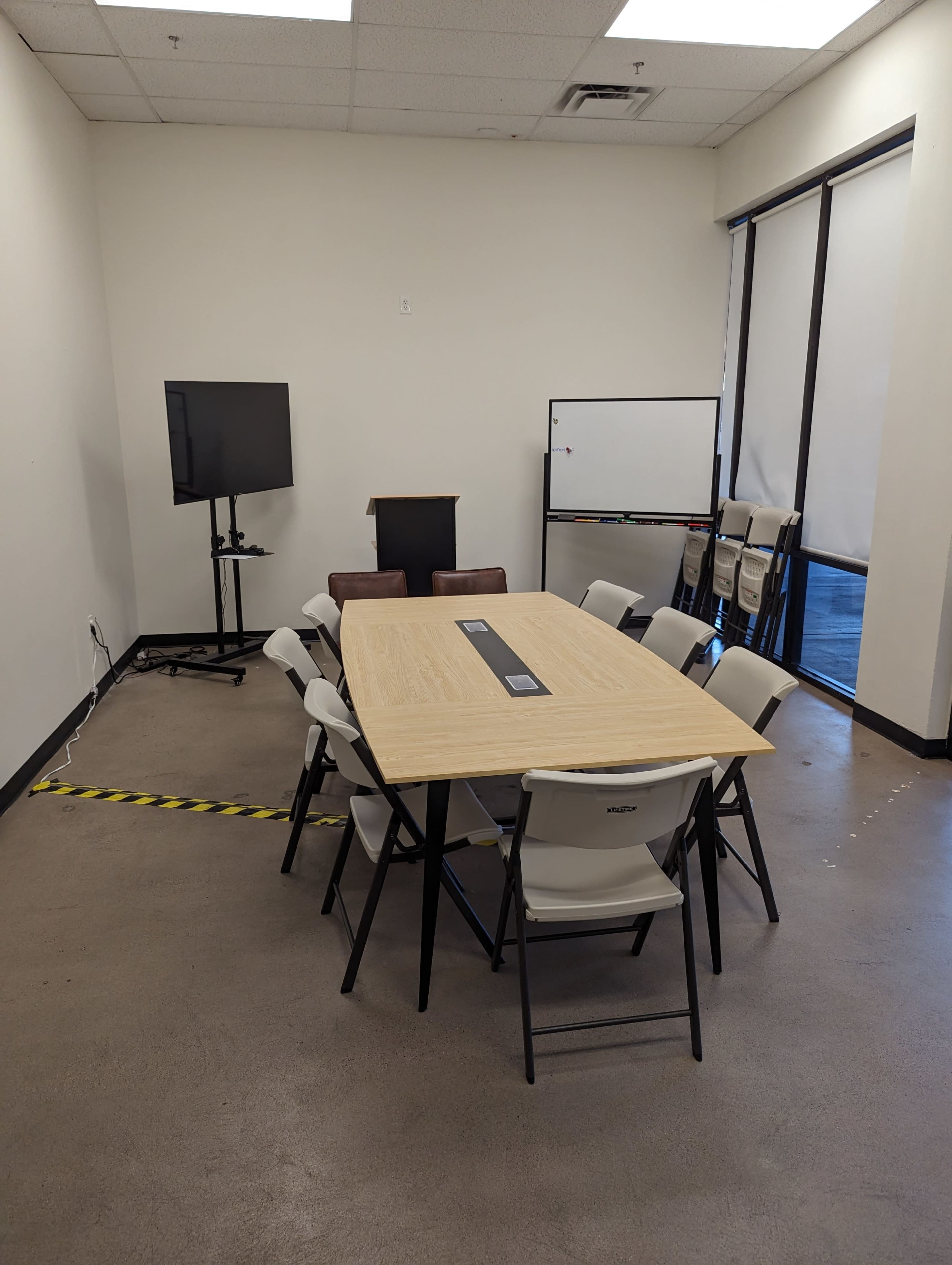 A conference room features a large wooden table surrounded by eight folding chairs, with a television on a stand and a whiteboard nearby.