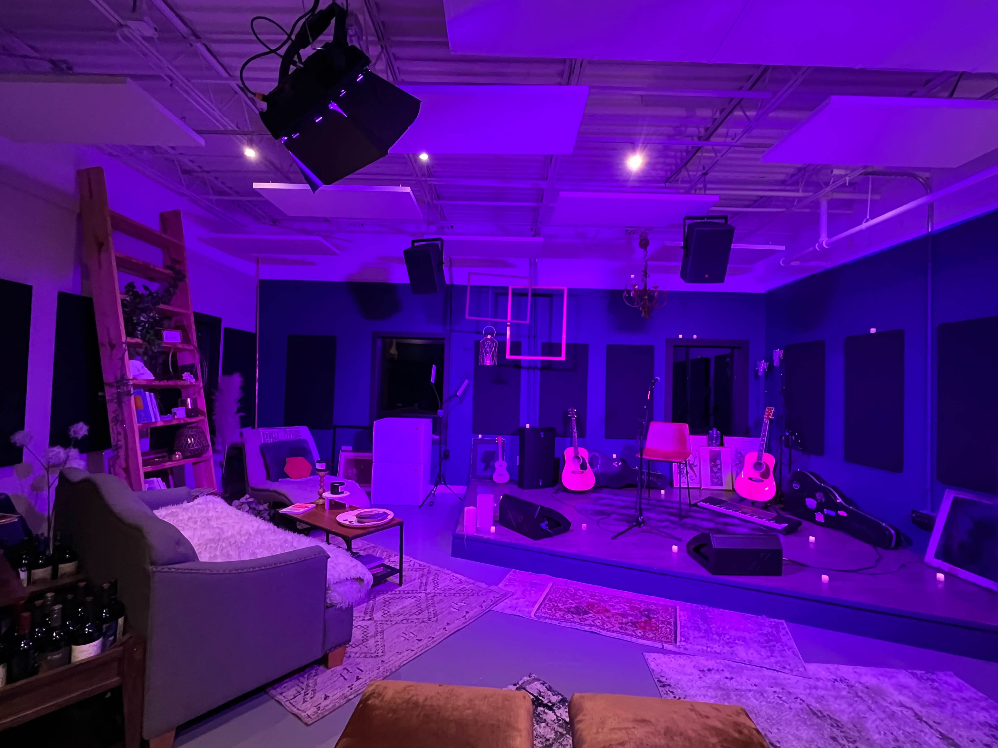 A music studio features a stage with guitars, a microphone, and audio equipment, complemented by a cozy seating area and purple lighting.