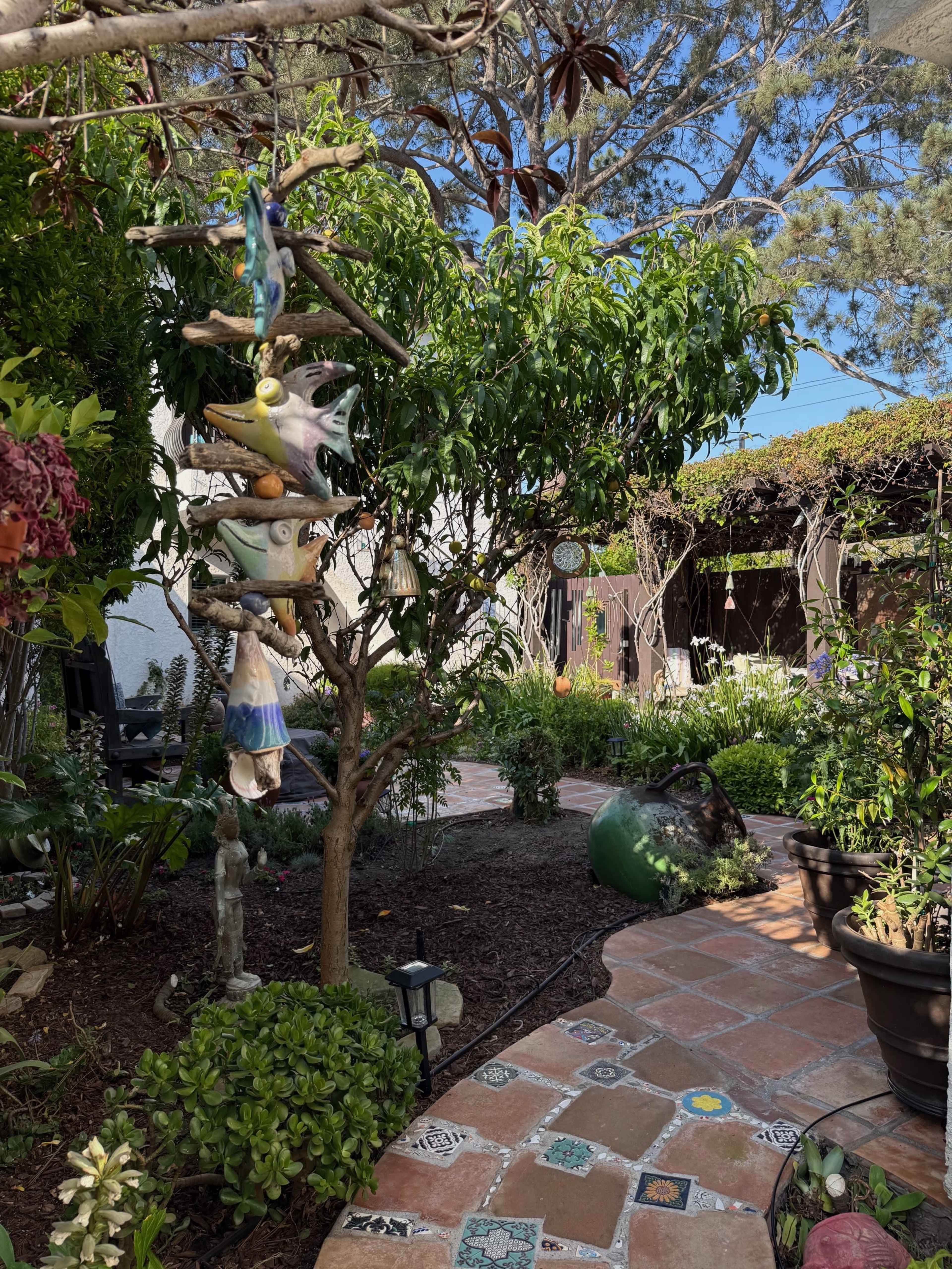 The image depicts a garden with a stone pathway, surrounded by various plants, trees, and decorative elements, including a hanging sculpture.