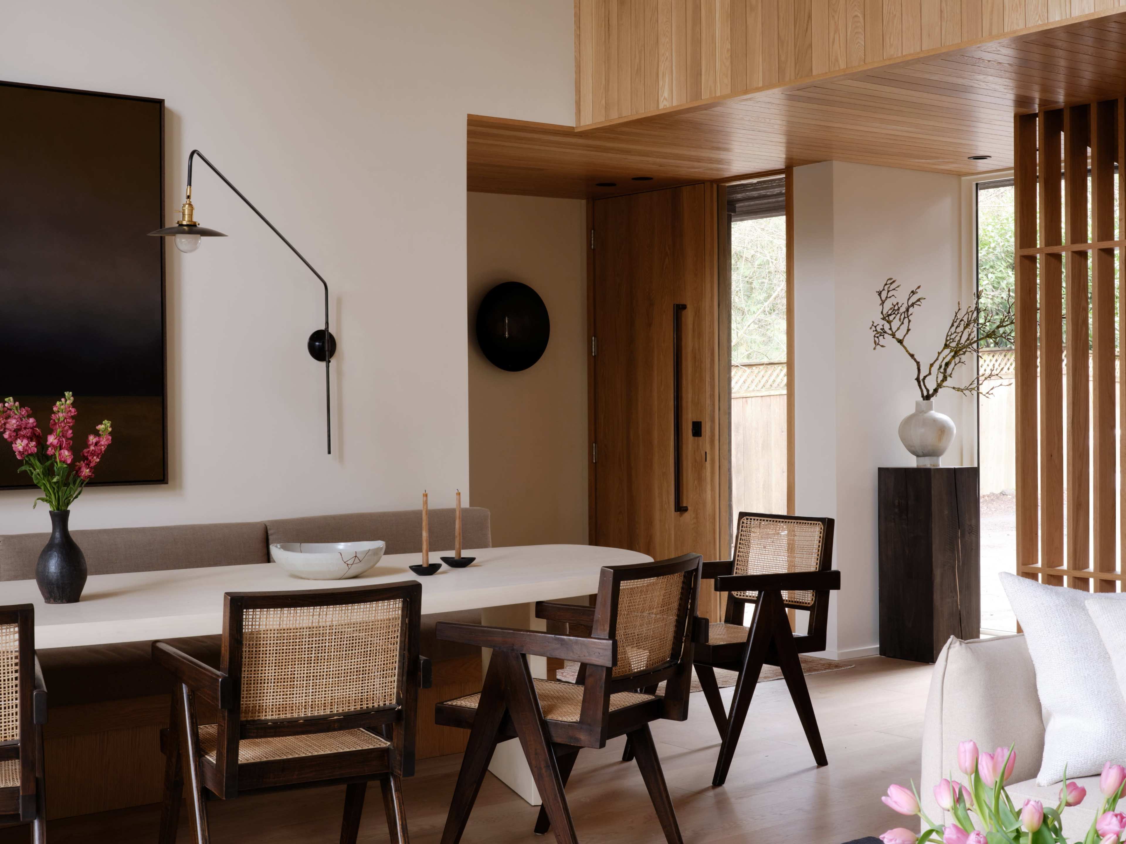 A modern dining area features a long, light-colored table surrounded by wooden chairs, with a large painting on the wall and decorative elements on display.