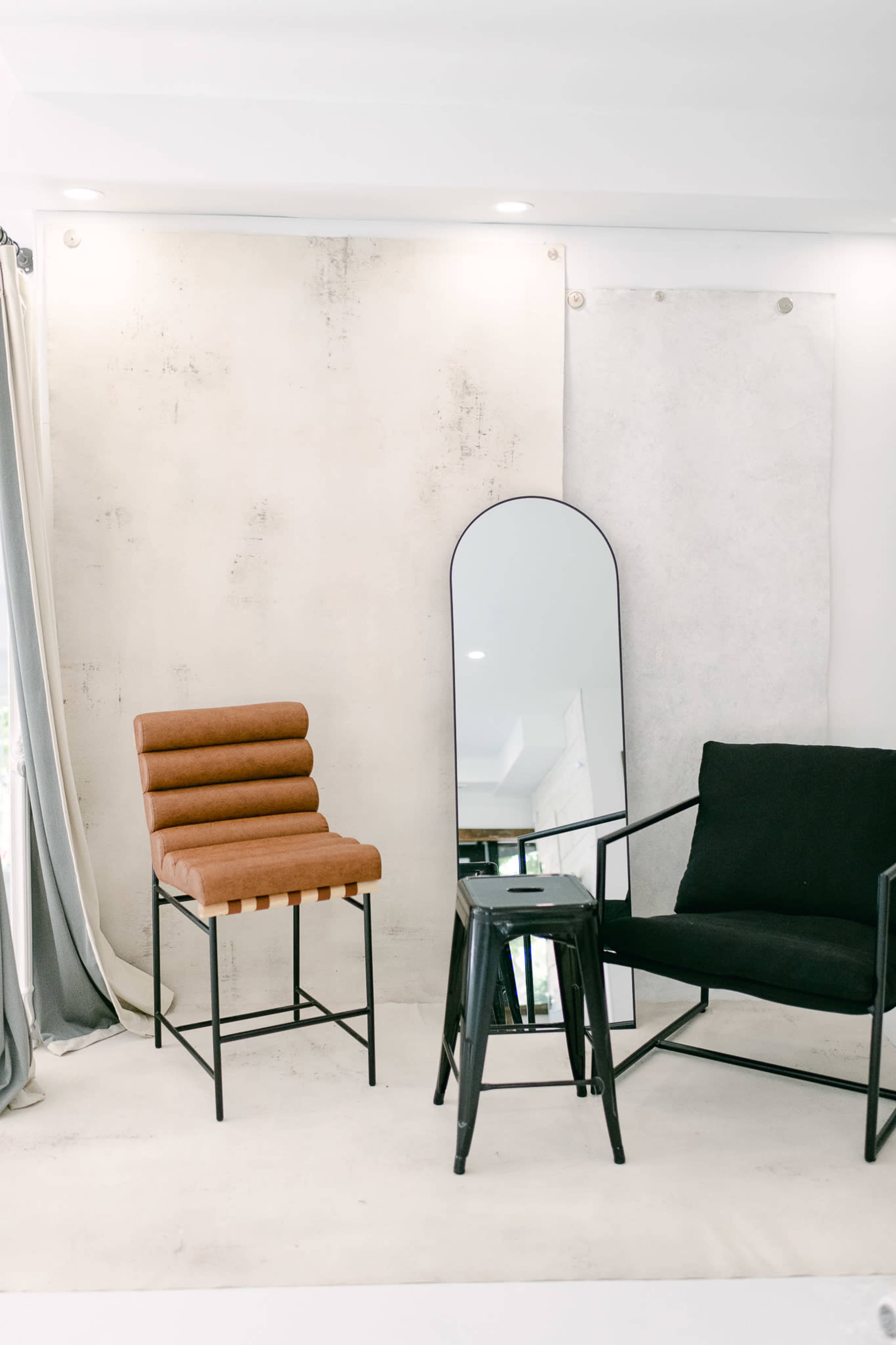 A minimalist interior space with a wooden bar stool, a black armchair, and a full-length mirror against a simple backdrop.