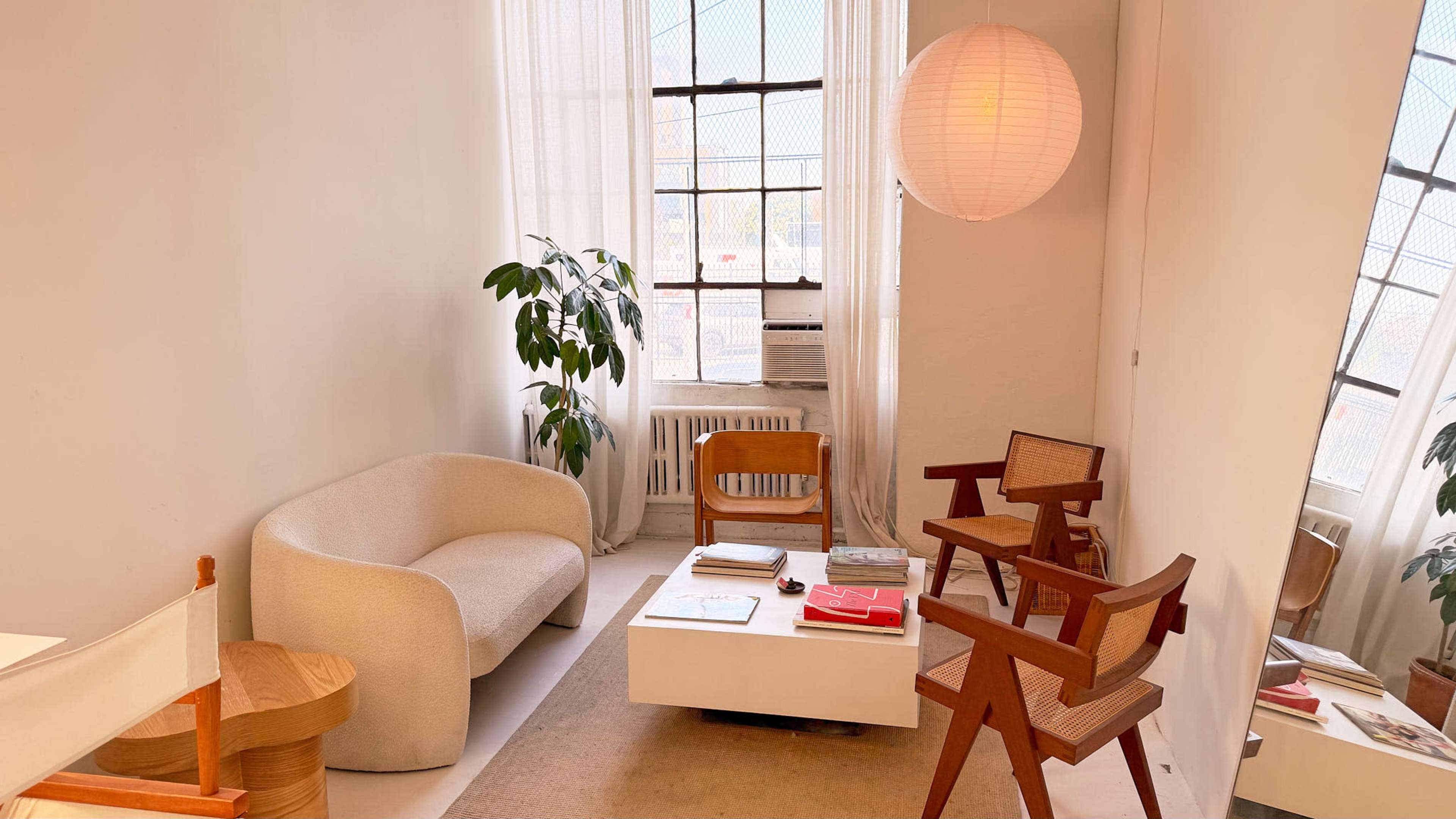 The image depicts a modern living area featuring a white sofa, a wooden chair, a round paper lantern light, and a coffee table, all set against a backdrop of large windows with sheer curtains.
