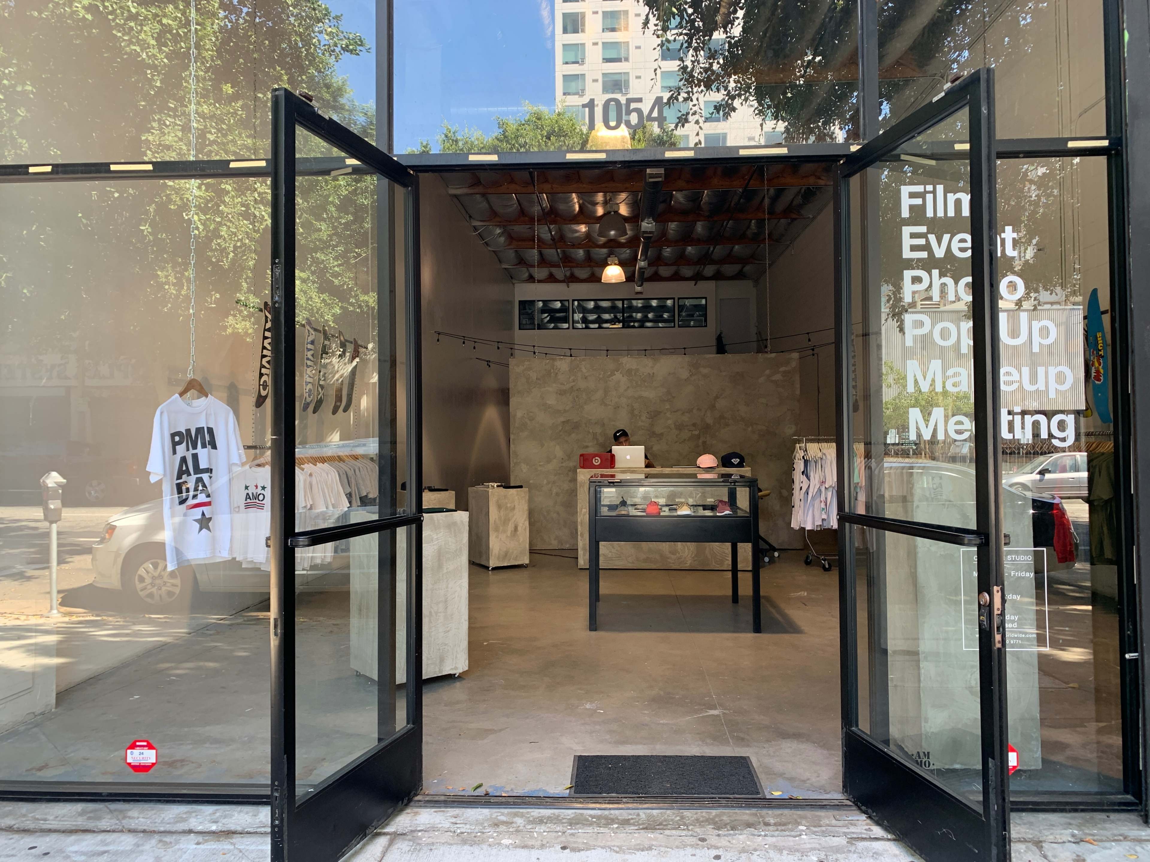 The image shows a storefront entrance with glass doors open, revealing a minimalist interior displaying clothing and a display case.