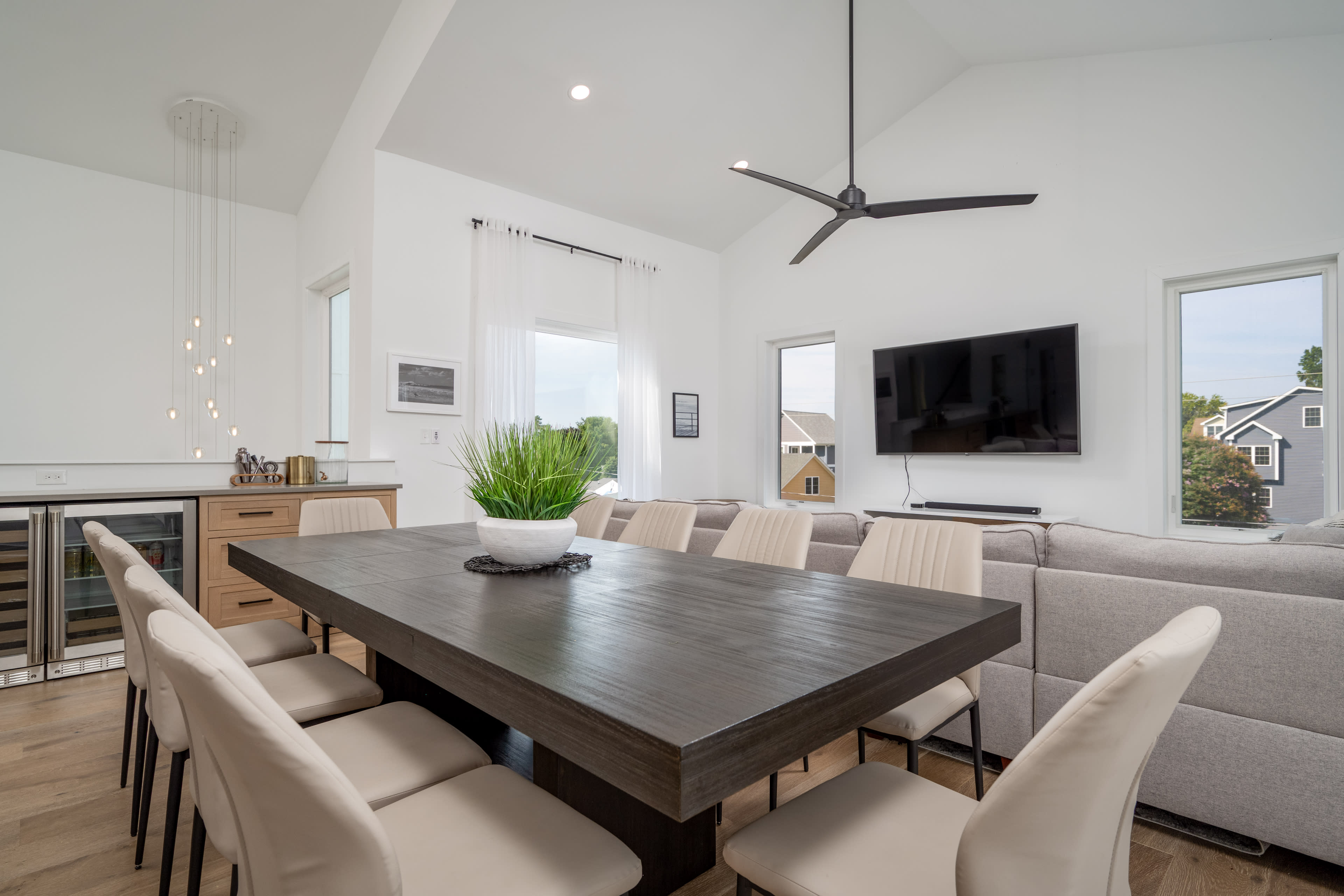 A modern dining area with a large wooden table surrounded by beige chairs, a ceiling fan, and a wall-mounted television.