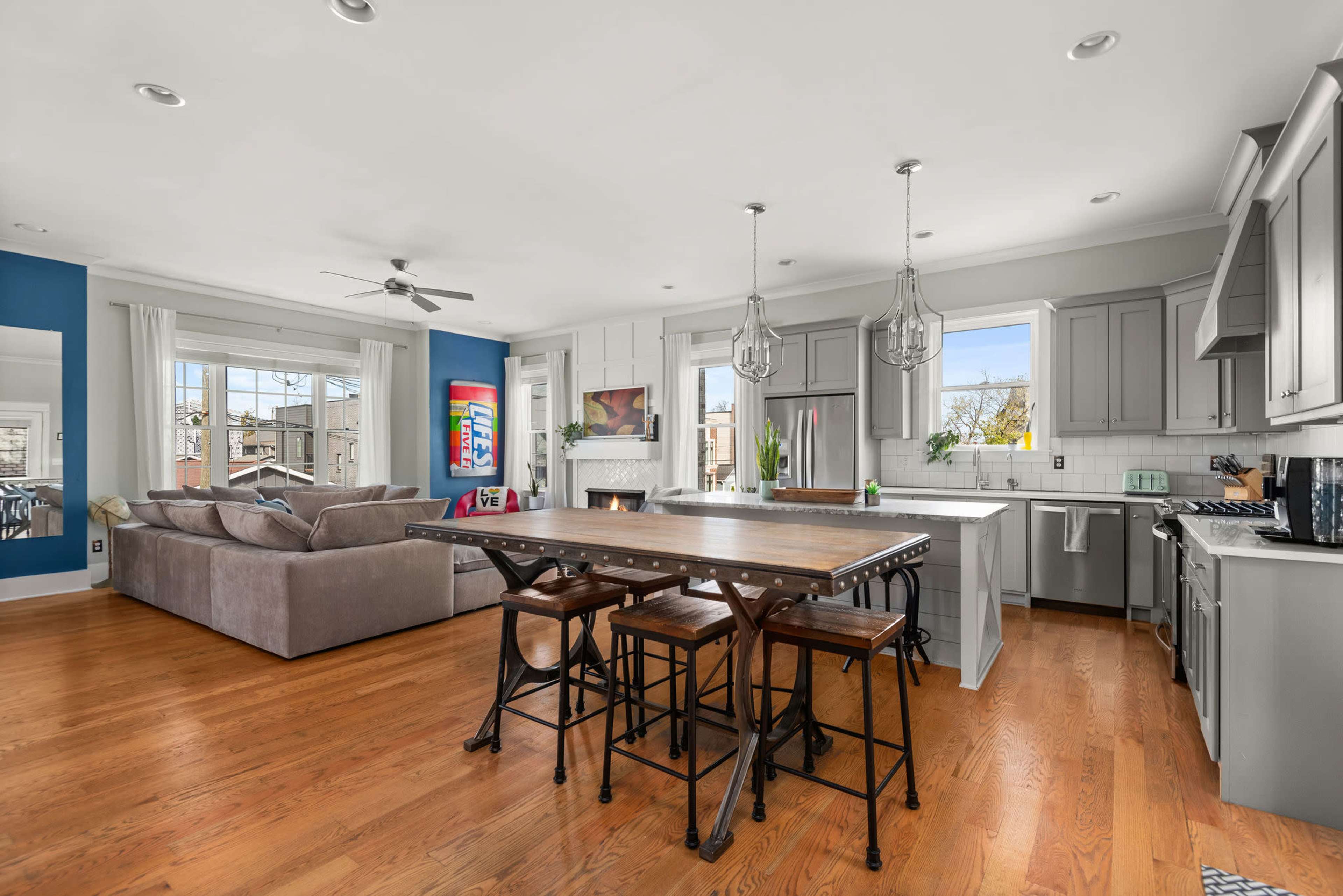 The image shows a modern, open-concept kitchen and living area with a large wooden dining table, gray cabinetry, and a comfortable sectional sofa.