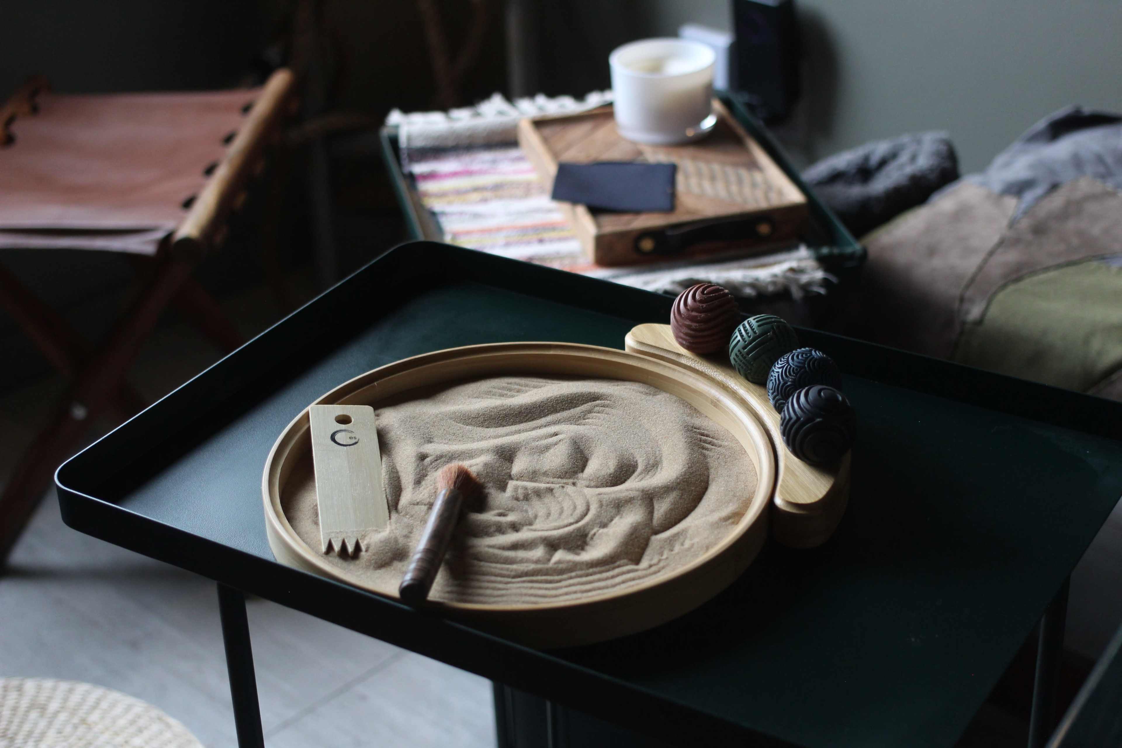 A circular wooden tray filled with fine sand, accompanied by a brush and three colored stones, sits on a dark table next to a folded chair and a decorative box.